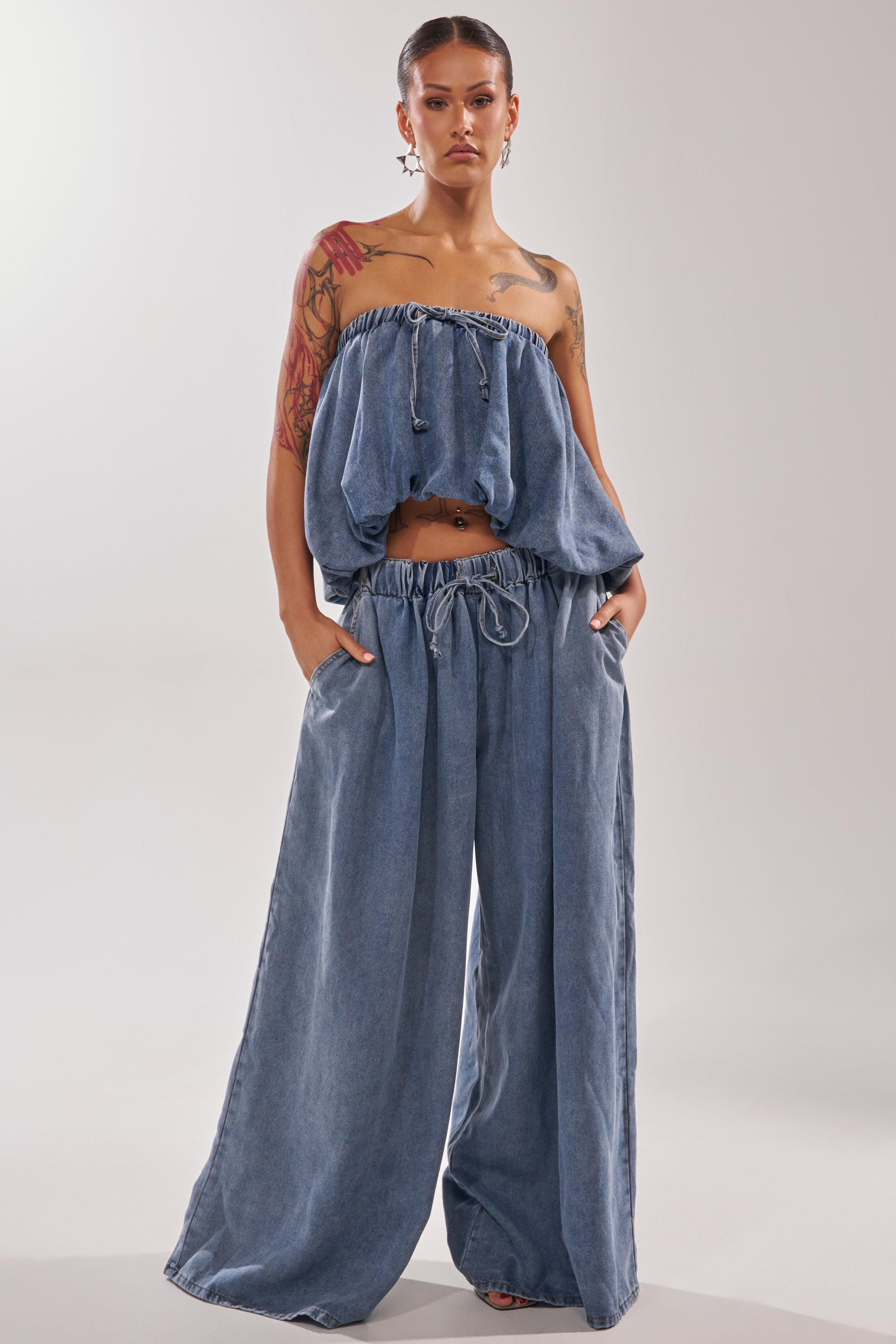 A person poses confidently in the DENIM FOR DAYS JOGGER, featuring high-rise, wide-leg denim pants with drawstring details and a loose, off-shoulder top. Tattoos complete their look against a simple light background. 5