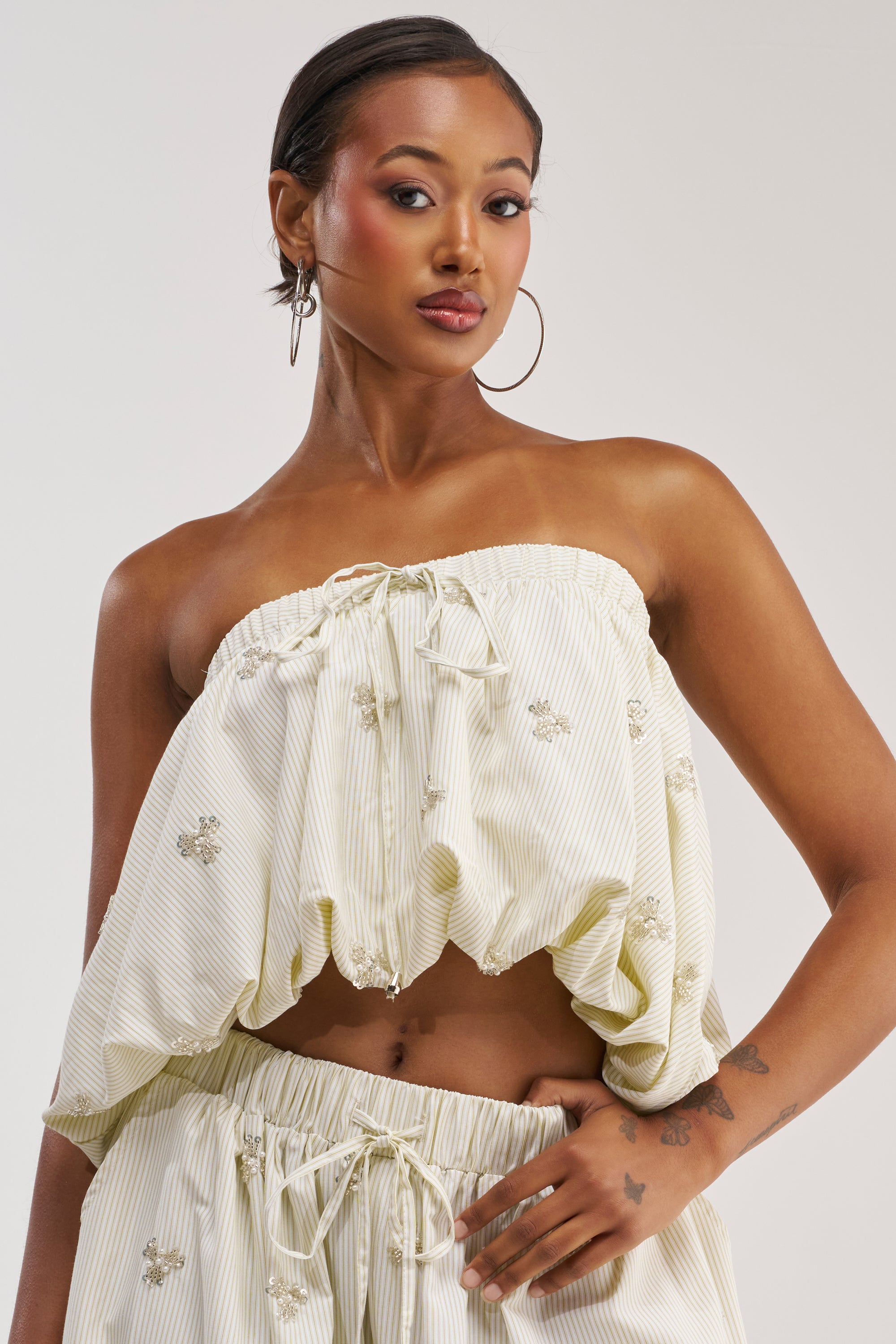 Dressed in the LIGHT AND AIRY POPLIN BLOUSE—an off-white, strapless design with ruffled accents and delicate floral embroidery—she completes her modern summer look with coordinating pants, hoop earrings, slicked-back hair, and a forearm tattoo.