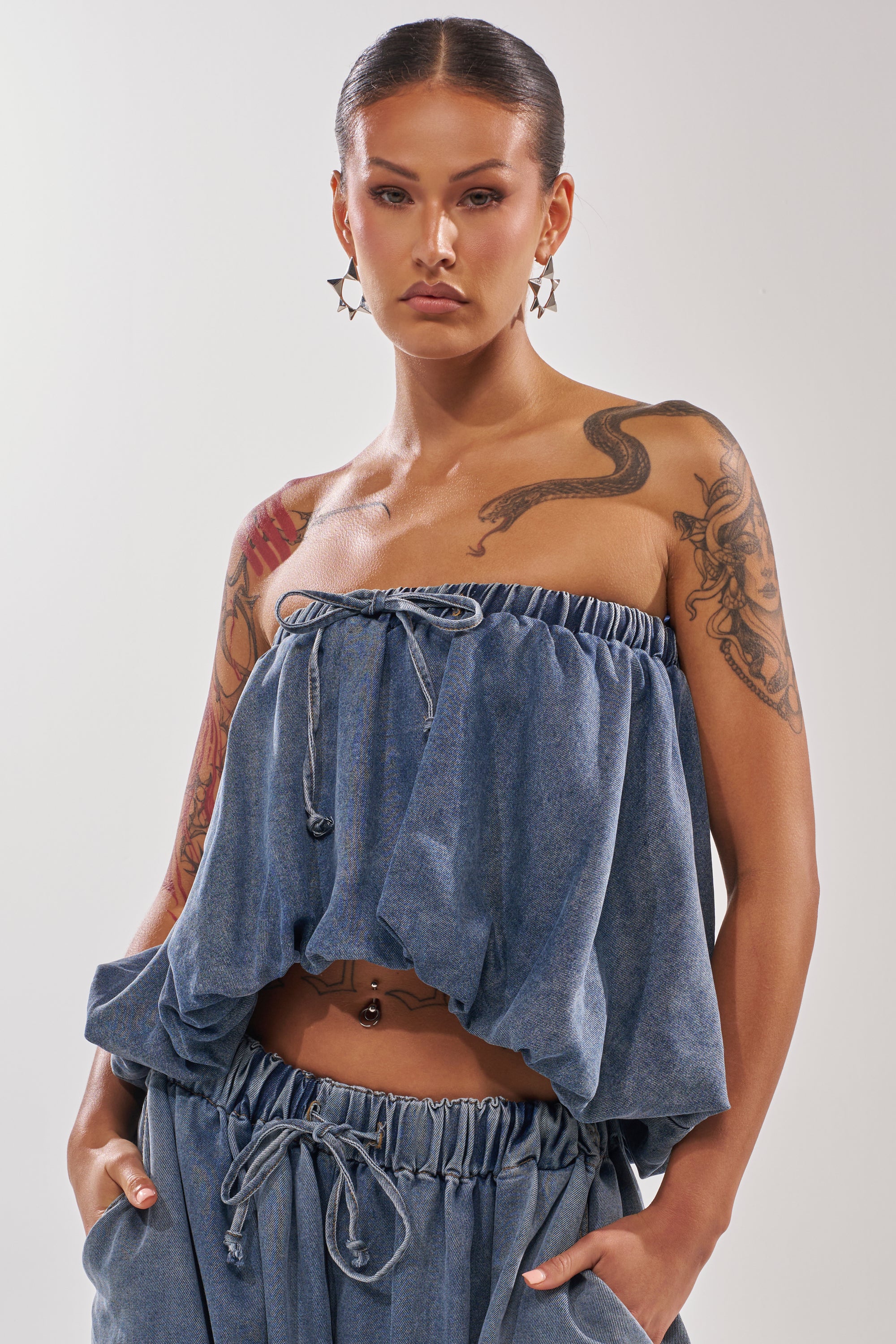 A tattooed woman models the DENIM FOR DAYS TUBE TOP—a chic strapless denim piece with a drawstring—paired with loose denim pants, star earrings, and slicked-back hair against a plain background. Hands are in her pockets. 0