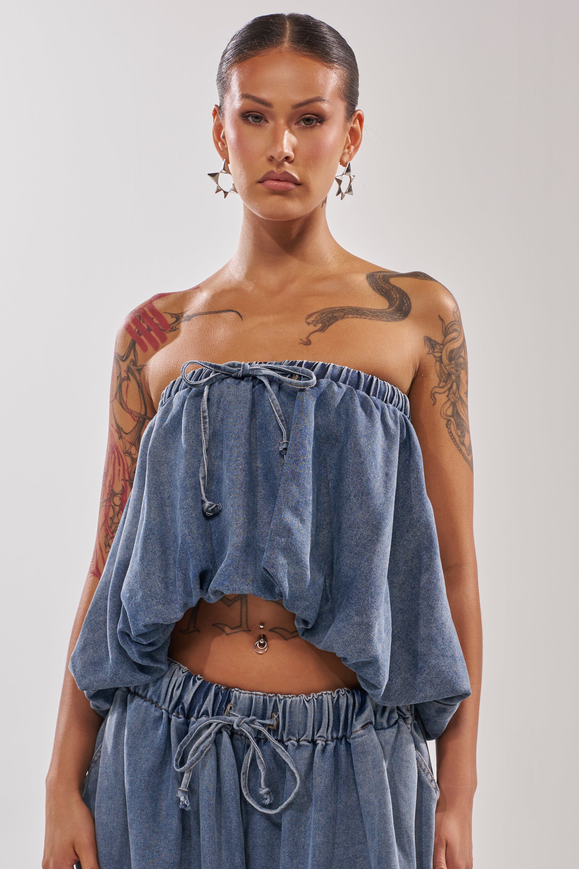 A woman with long hair and hoop earrings wears the DENIM FOR DAYS TUBE TOP with matching drawstring pants. Her arm, shoulder, and chest tattoos and belly button piercing complete her bold summer-chic look. 1