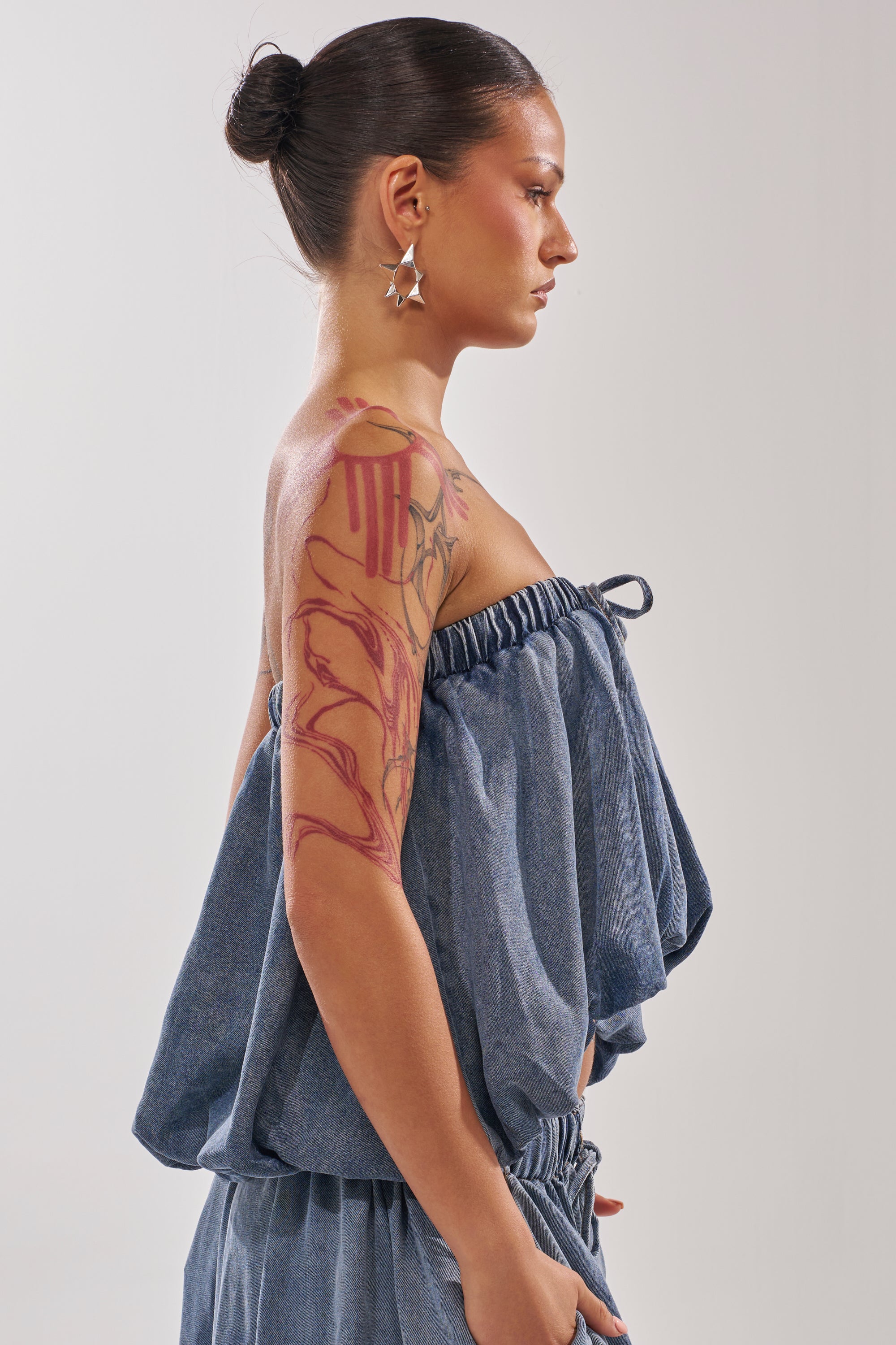 A woman with dark hair in a bun, wearing star-shaped earrings and the DENIM FOR DAYS TUBE TOP with matching pants, stands in profile. A large artistic red tattoo covers her upper right arm and shoulder. 2