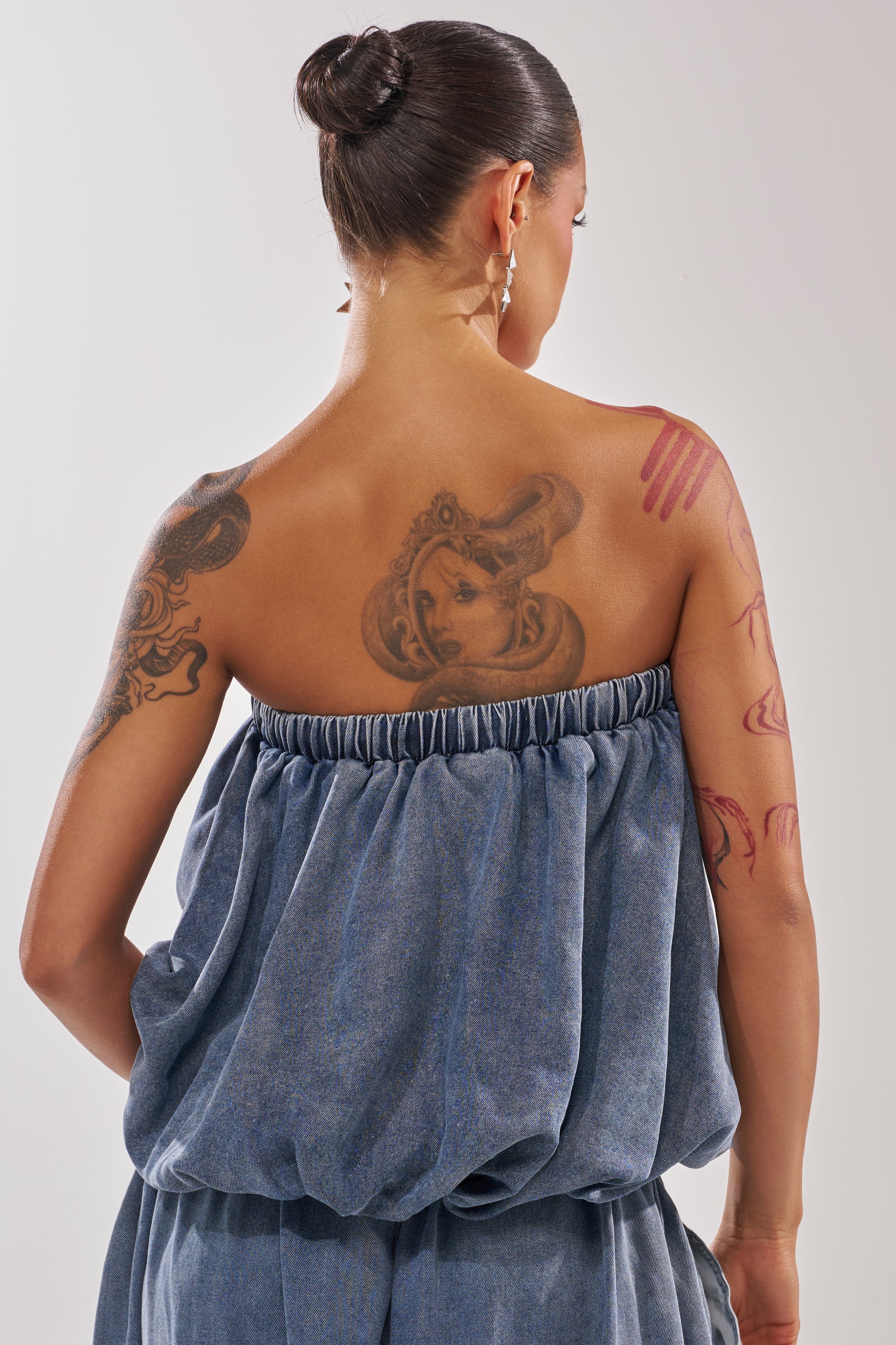 A woman with her back to the camera showcases detailed upper back and right arm tattoos, wearing the DENIM FOR DAYS TUBE TOP. Her dark hair is styled in a sleek bun. 3