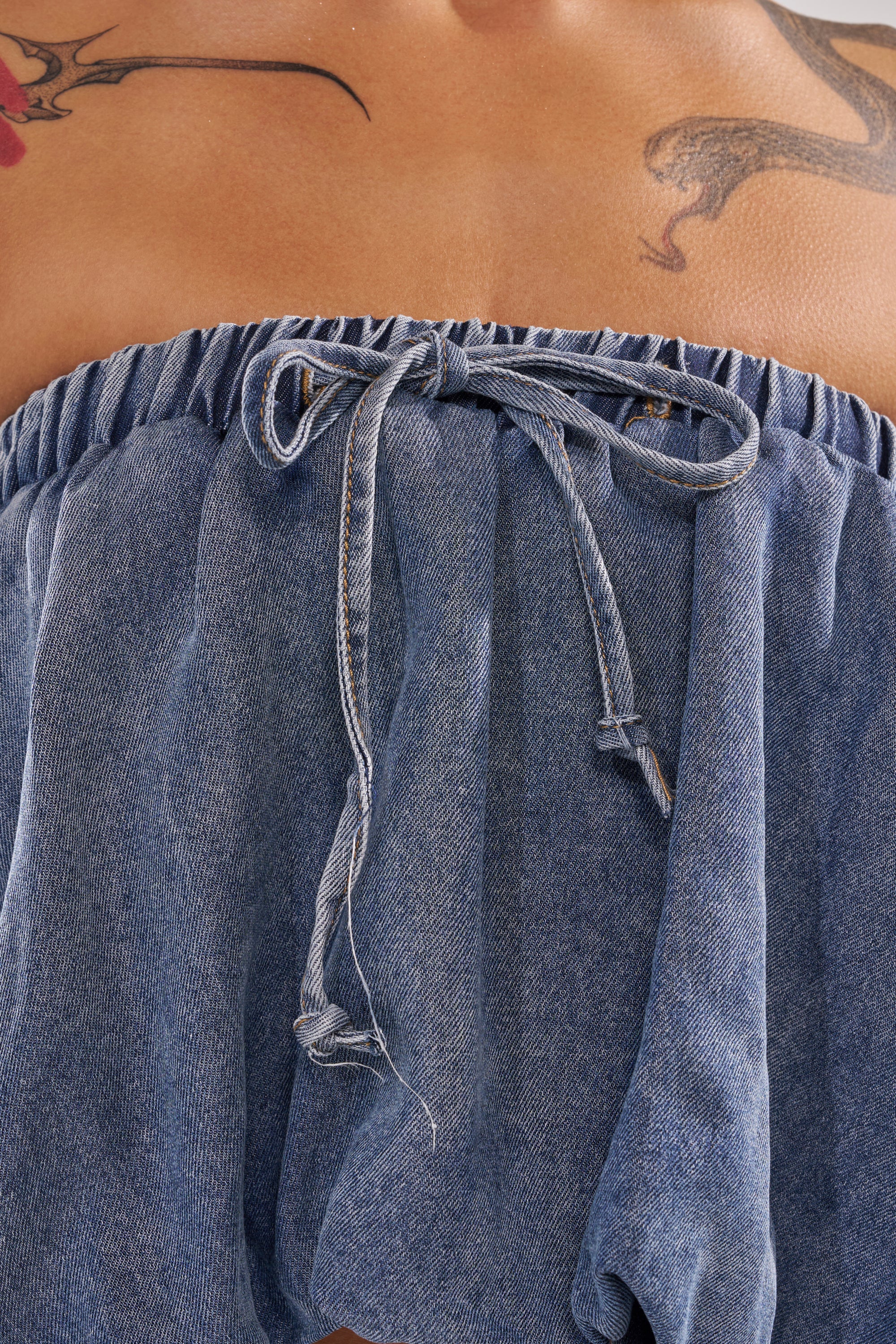 Close-up of a person wearing the DENIM FOR DAYS TUBE TOP, a strapless denim style with a center bow. Visible upper chest and shoulder tattoos—a red flower and dragon—add edge to this summer-chic look. 7