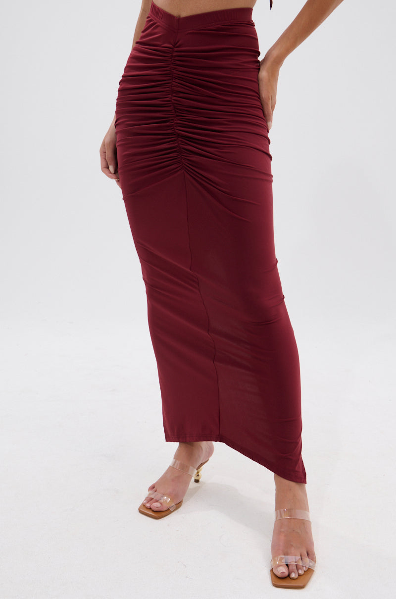 A person wears the SEXY RUCHED BACK SLIT MAXI SKIRT in burgundy with clear strap heeled sandals, standing against a plain white background. The high-rise skirt features gathered details down the center front for a flattering effect. 3