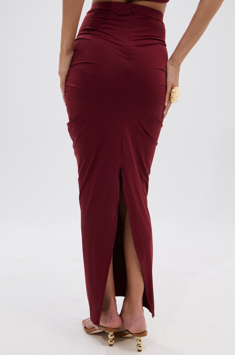 A person is seen from behind in the SEXY RUCHED BACK SLIT MAXI SKIRT—a high-rise, ankle-length burgundy skirt with a ruched design and back slit—paired with gold-accented heels, set against a plain white background. 5