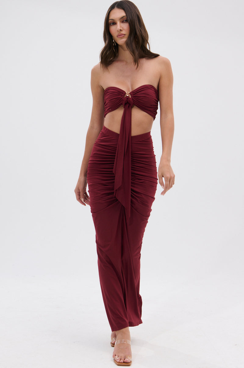 A woman models the SEXY RUCHED BACK SLIT MAXI SKIRT in burgundy, featuring a strapless, ruched design with a front knot and midriff cutout, paired with clear heels against a plain white background. 7