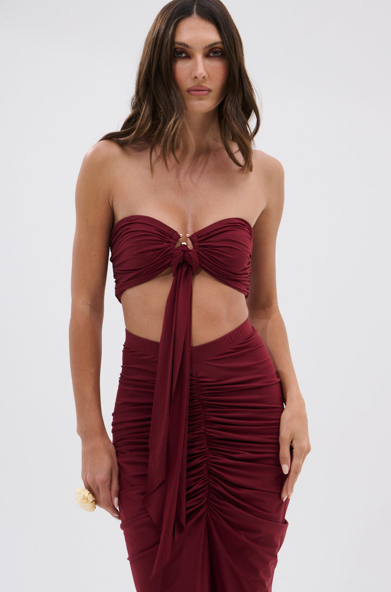 A woman models the SEXY RUCHED BANDEAU TOP in burgundy, featuring a front tie, paired with a matching high-waisted ruched skirt, set against a plain light background. 1
