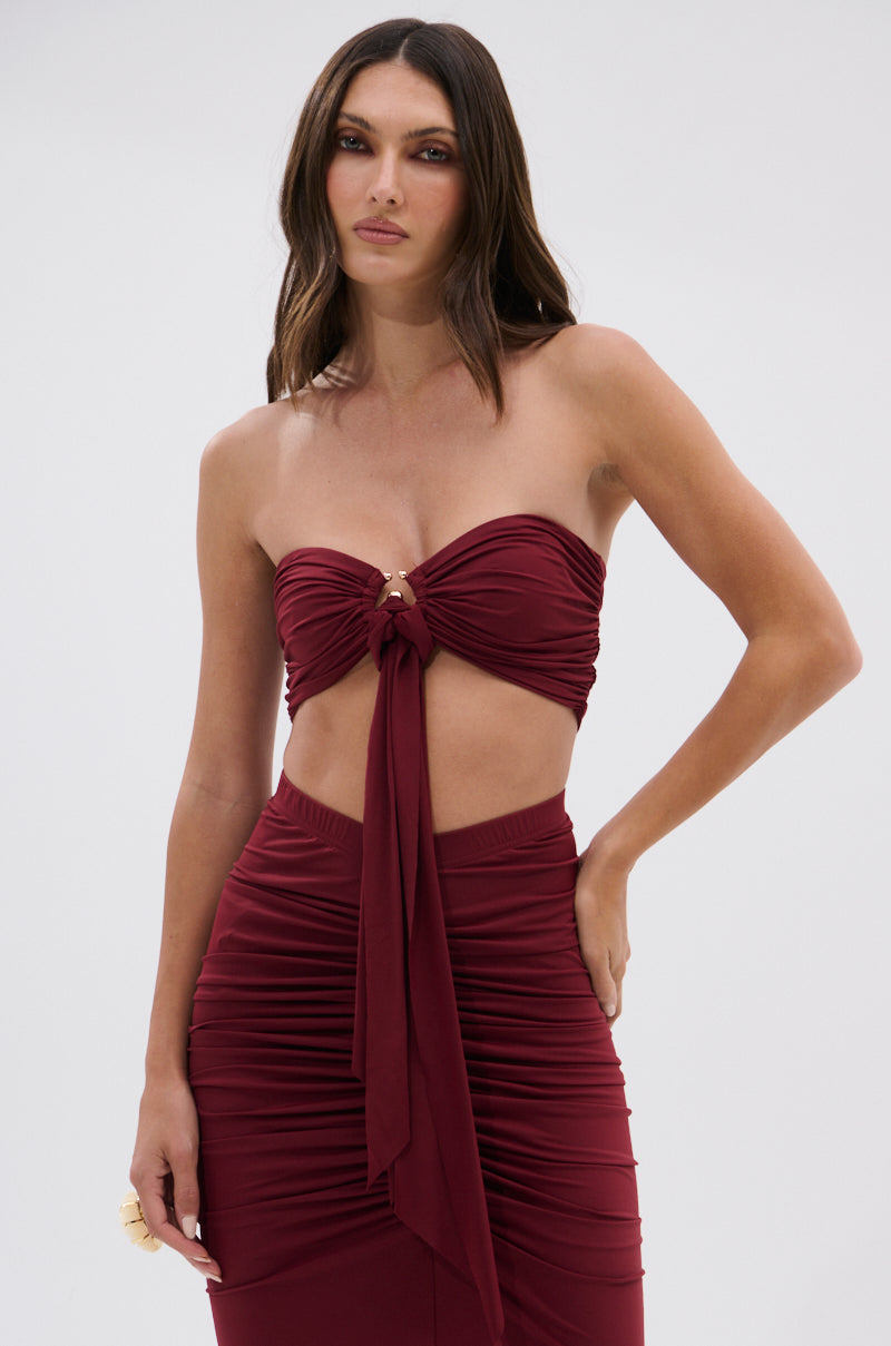 A woman models the SEXY RUCHED BANDEAU TOP, a strapless burgundy piece with a gold O-ring centerpiece and midriff cutout, standing against a plain light background. 2