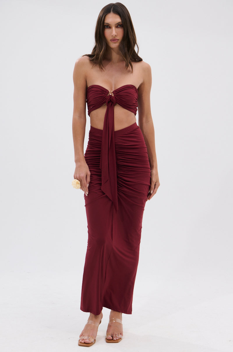 A woman wears the SEXY RUCHED BANDEAU TOP dress in dark red with a front cutout and gold o-ring detail, styled with clear high-heeled sandals and straight brown hair, against a plain white background. 5