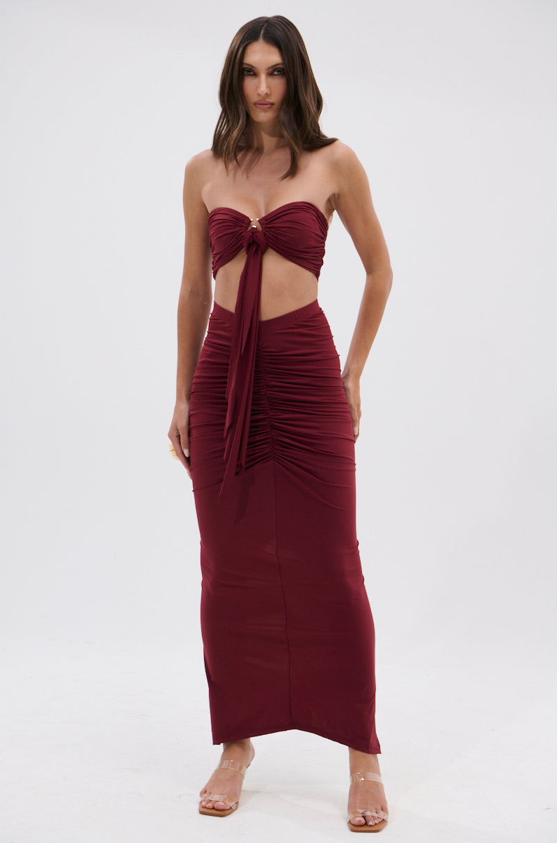 A woman stands facing forward against a plain white background, wearing the SEXY RUCHED BANDEAU TOP in dark red with a front tie and midriff cutout, paired with a matching maxi skirt. She has loose wavy hair and clear high-heeled sandals. 6