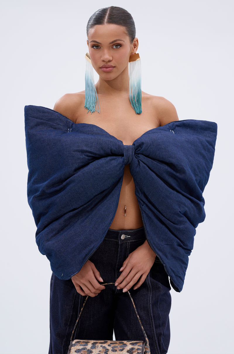 A woman stands against a plain light background wearing the PADDED DENIM BOW TOP with a sweetheart neckline, dark denim pants, ombre blue-and-white fringe earrings, and holding a small leopard print bag.