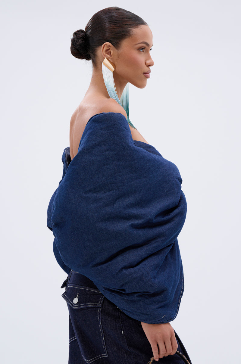 A woman with dark hair in a bun models the PADDED DENIM BOW TOP, styled with long blue gradient earrings and a matching blue skirt, posing in profile against a plain white background.