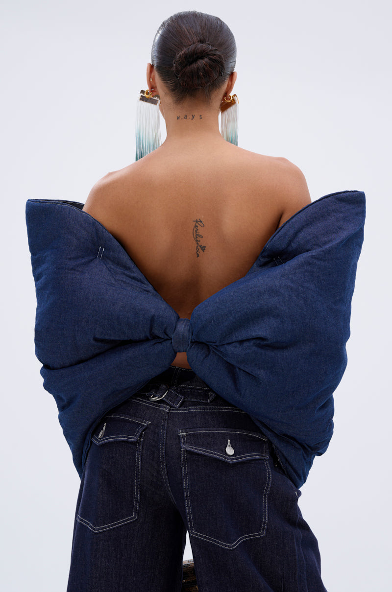 A person with their back to the camera models the PADDED DENIM BOW TOP—an oversized bow silhouette worn over dark-wash jeans. With hair in a bun, they display earrings and back tattoos against a plain background.