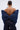 A person with their back to the camera models the PADDED DENIM BOW TOP—an oversized bow silhouette worn over dark-wash jeans. With hair in a bun, they display earrings and back tattoos against a plain background.