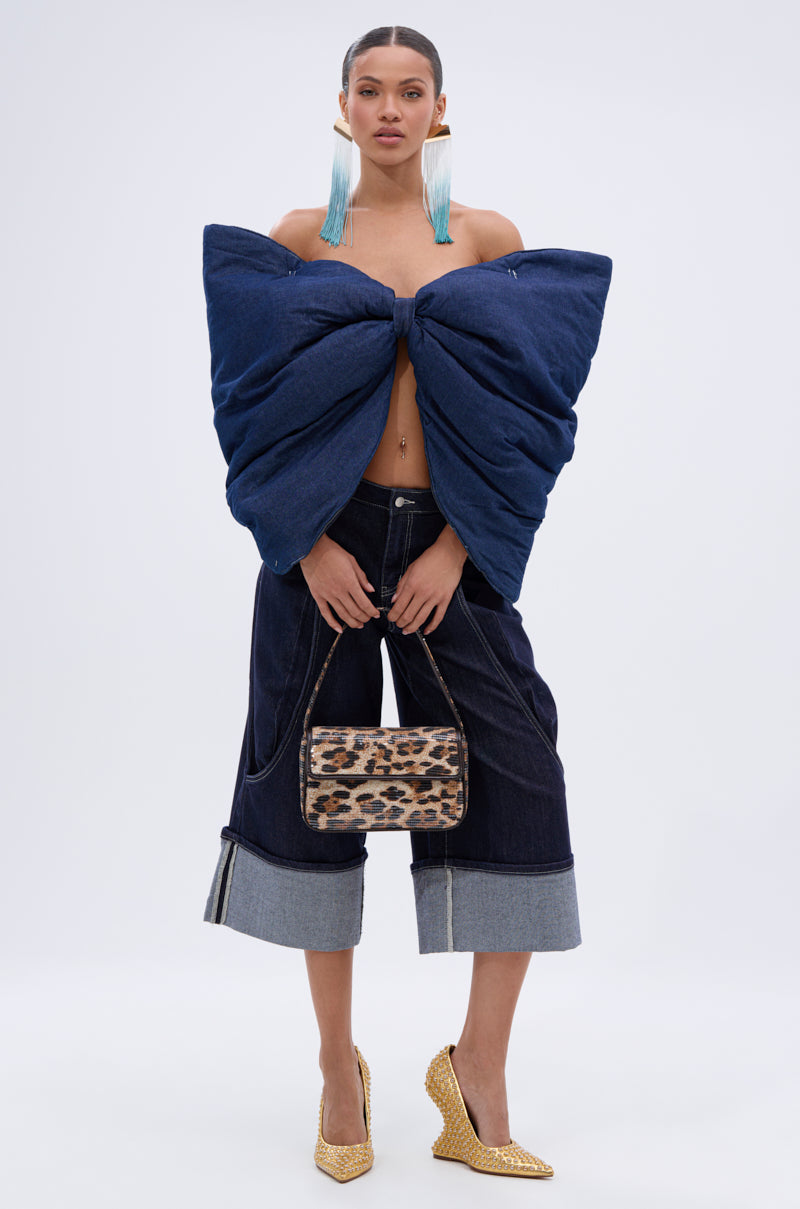 A woman poses in a PADDED DENIM BOW TOP with an oversized bow, paired with wide-leg cuffed dark-wash jeans, gold textured heels, blue tassel earrings, and a small leopard-print handbag.