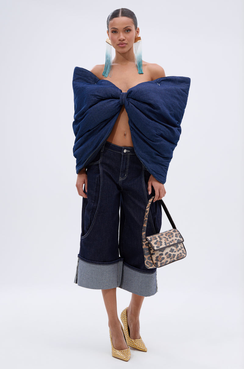 A woman poses against a plain light background wearing the PADDED DENIM BOW TOP with an oversized bow silhouette, paired with wide-leg cuffed dark-wash denim pants, yellow heels, blue statement earrings, and a leopard-print handbag.