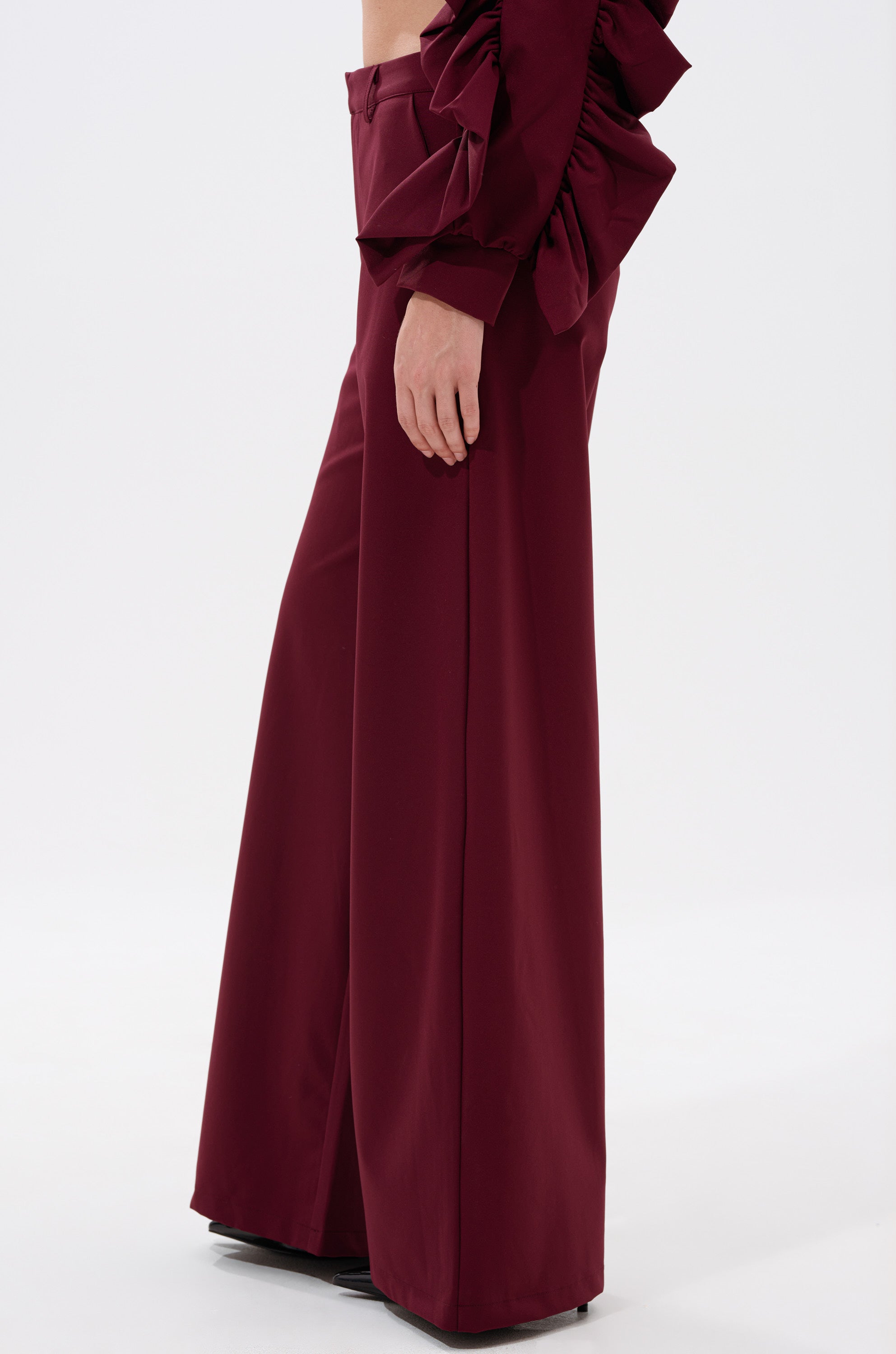 Someone is seen wearing the maroon SIERRA TROUSER, a high-rise, wide-leg pant, paired with a matching top with voluminous ruffled sleeves, posed against a plain white background. 2