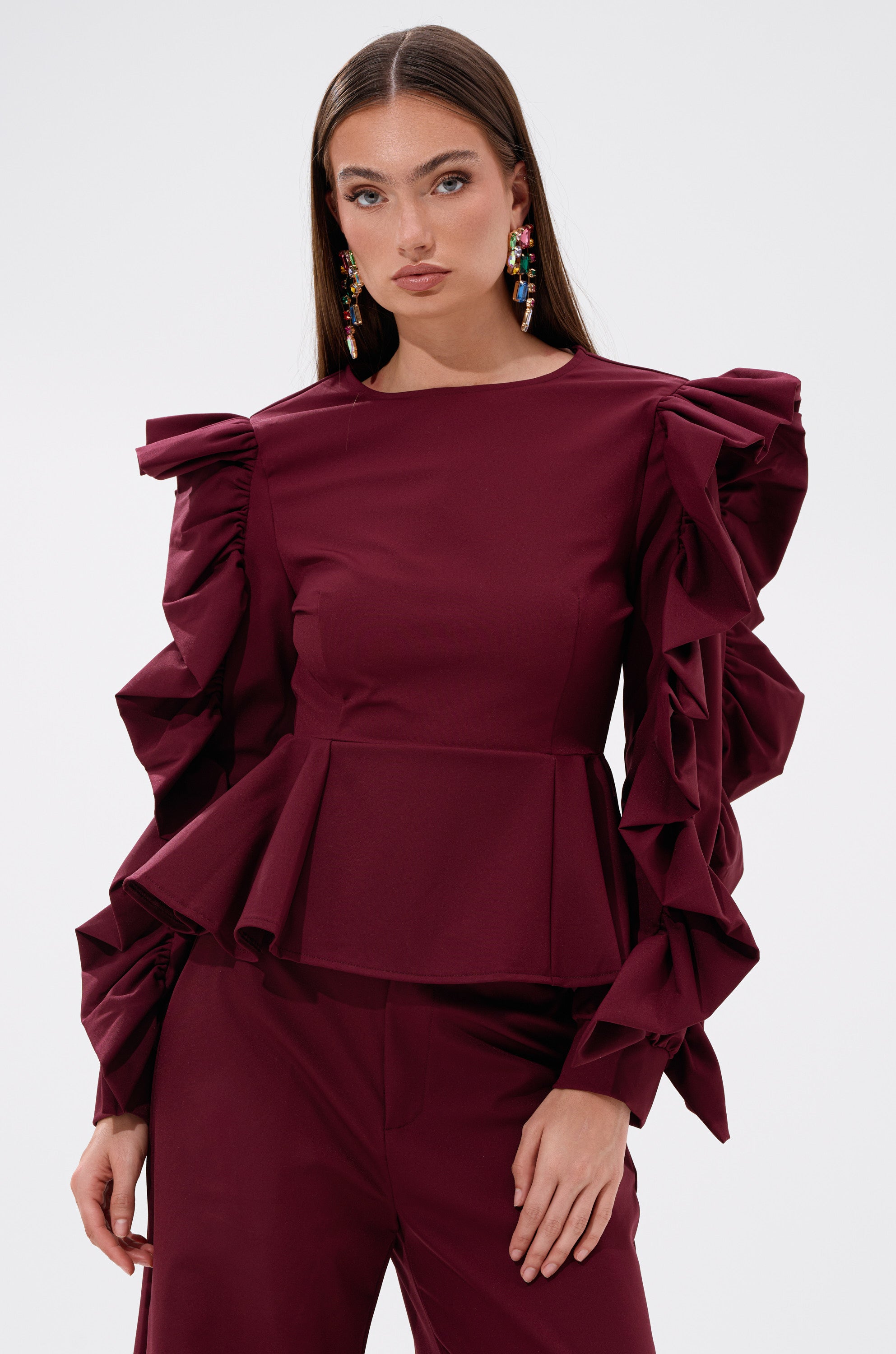A woman wears the SIERRA RUFFLE BLOUSE, which has a peplum hem and bold cascading ruffles, paired with matching pants. She stands against a plain white background with long straight hair, colorful earrings, and a neutral expression. 1