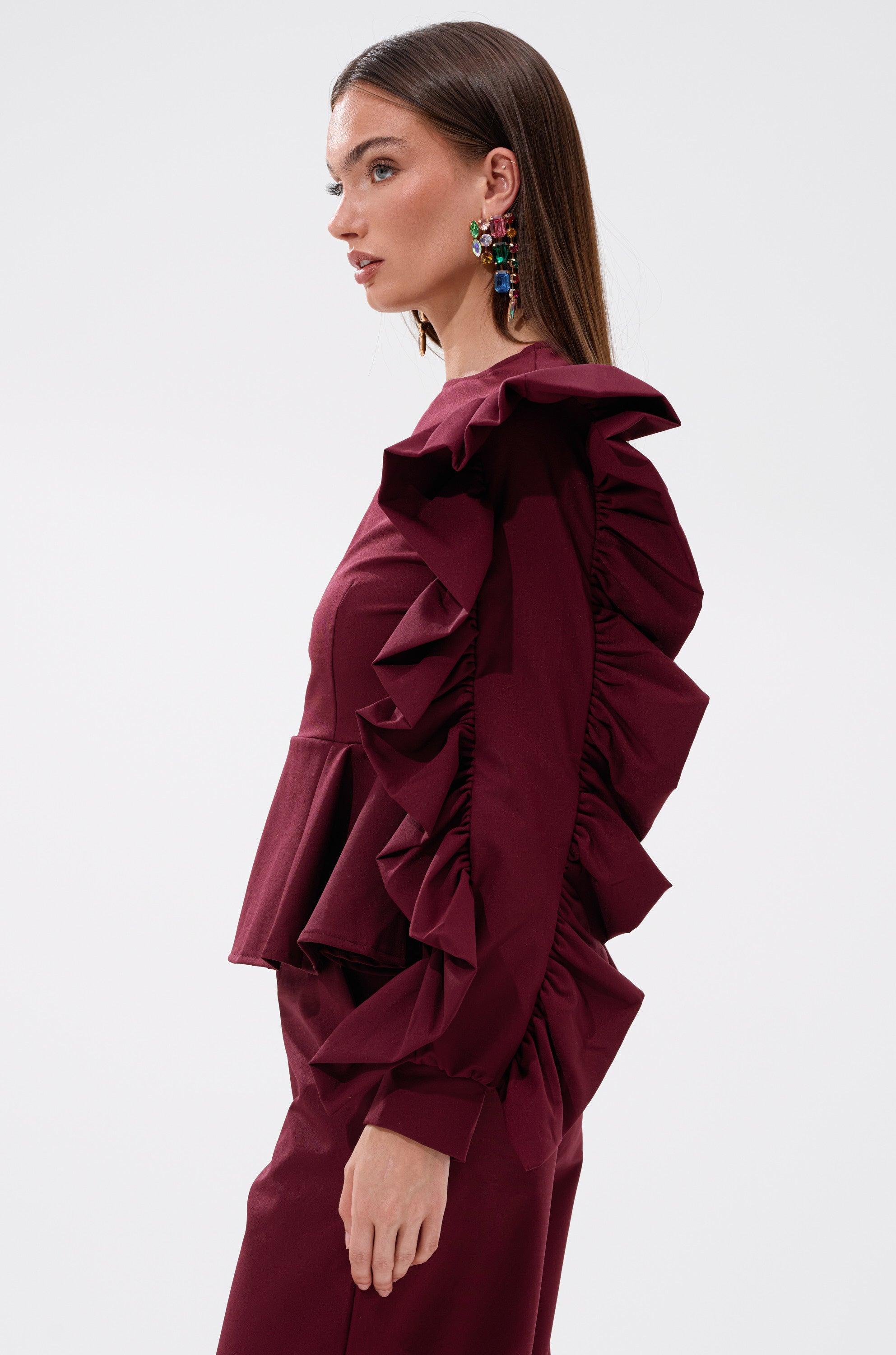 A woman with straight brown hair and colorful statement earrings models the SIERRA RUFFLE BLOUSE, featuring cascading ruffles on the sleeves and shoulders, paired with matching pants against a plain white background. 2