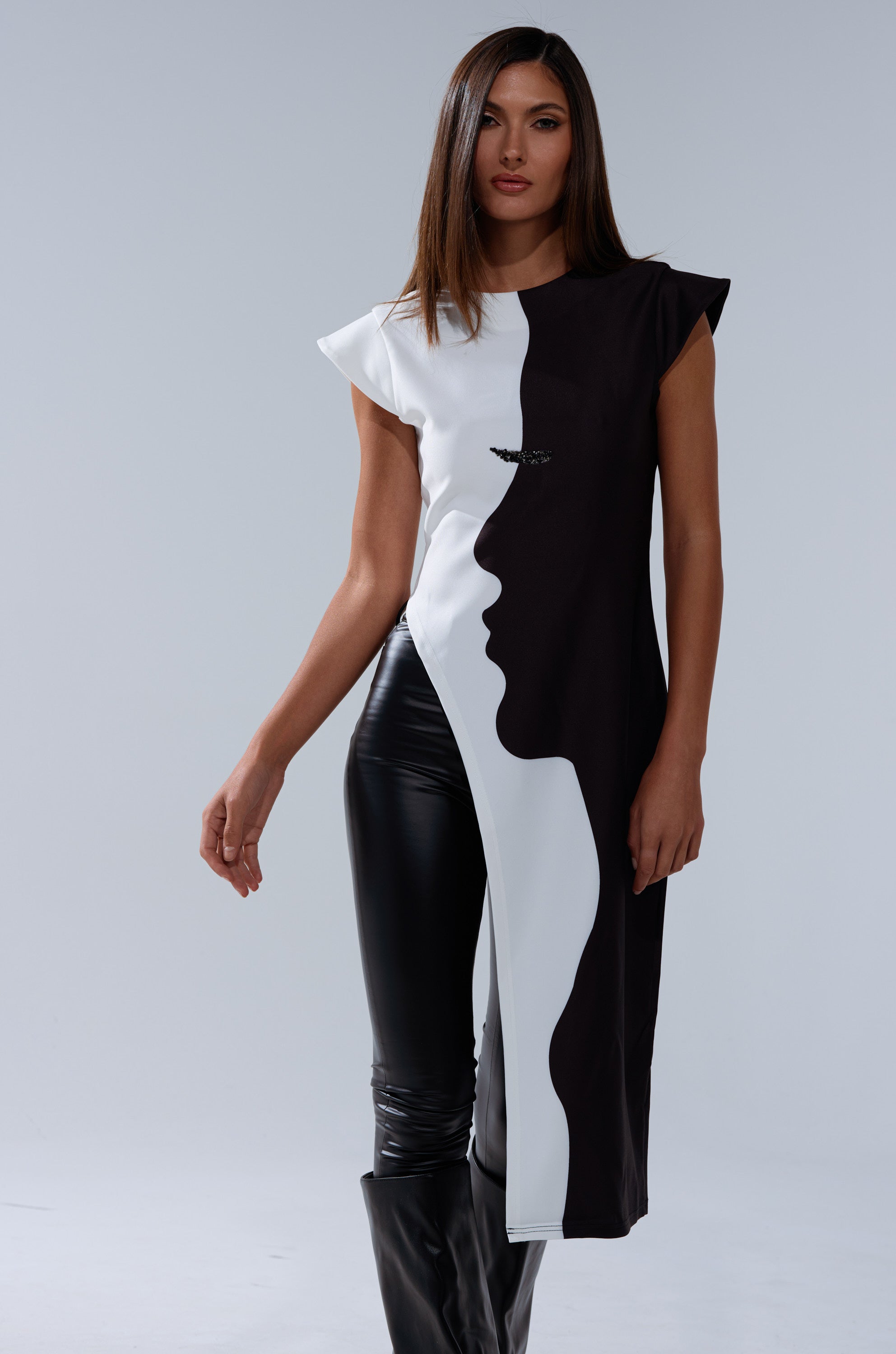 A woman with straight brown hair models the SILHOUETTE LONGLINE TEE, featuring an abstract black-and-white face graphic, paired with black leather pants against a plain light gray background.