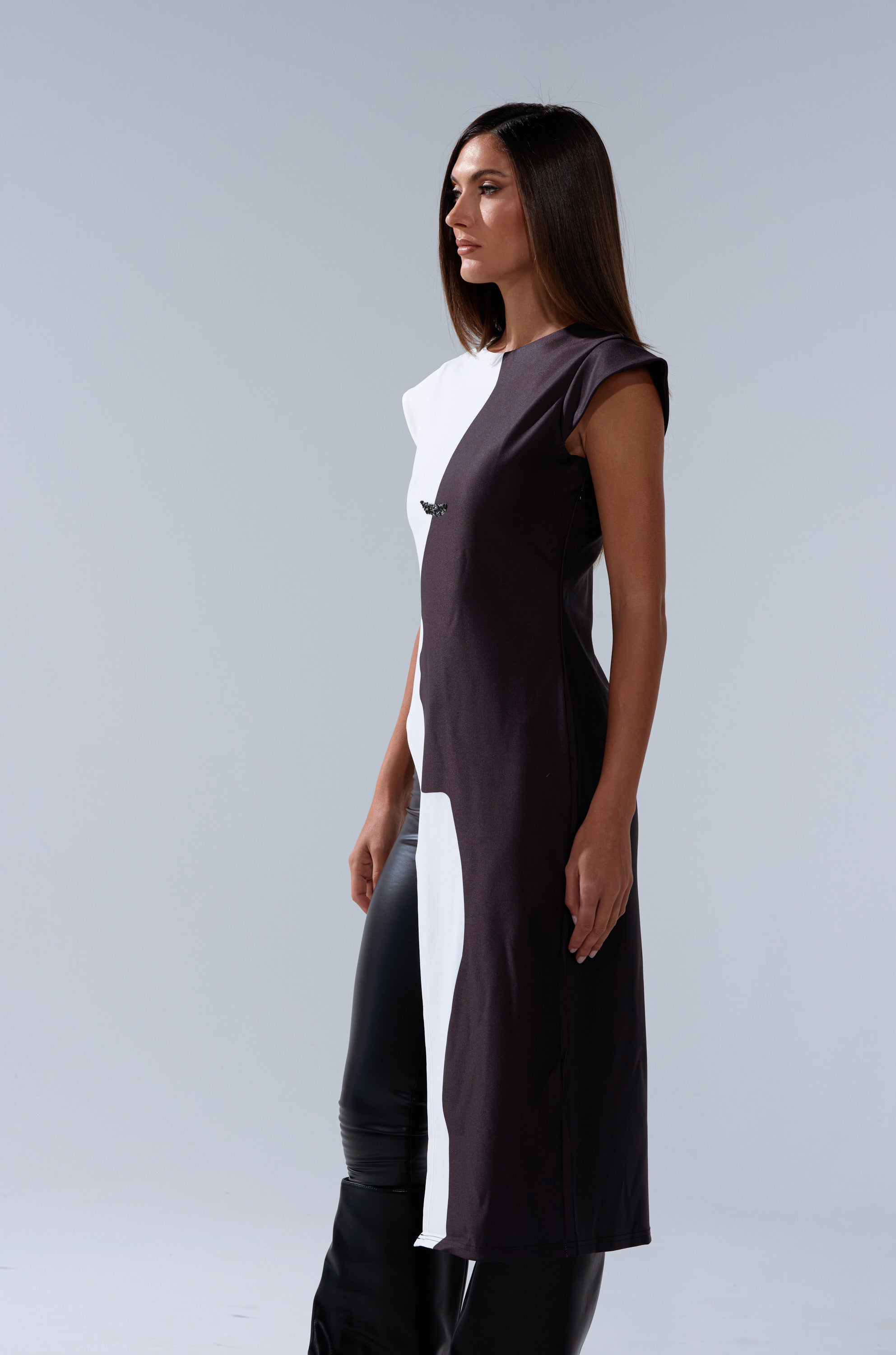 A woman with straight brown hair stands in a studio, looking to the side while wearing the SILHOUETTE LONGLINE TEE—a sleek, two-tone short-sleeve top paired with black pants—its bold color blocking contrasting against a light gray background.