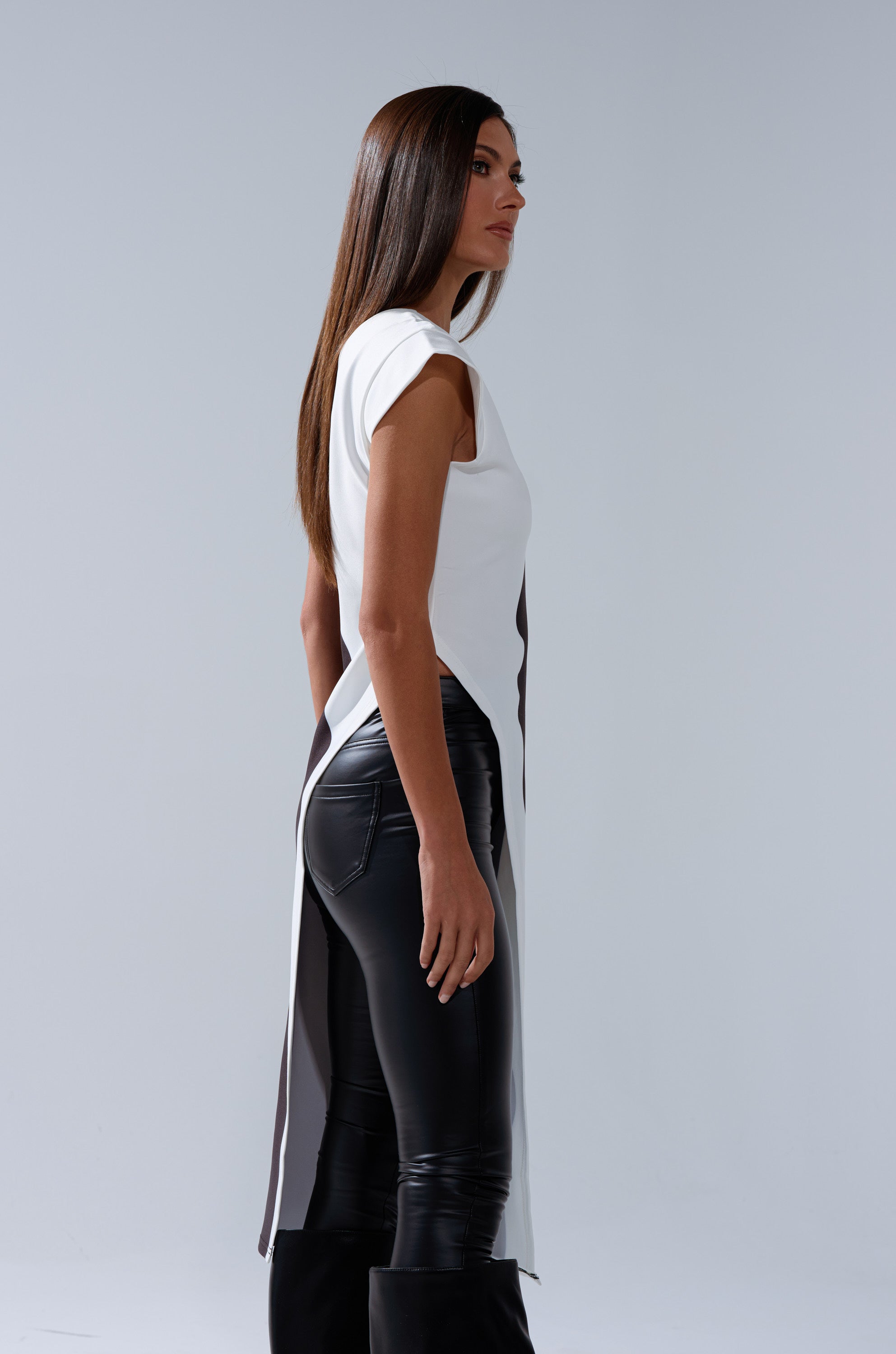 A woman with long, straight brown hair stands in profile, wearing the SILHOUETTE LONGLINE TEE over black leather pants and black boots, set against a plain light gray background.