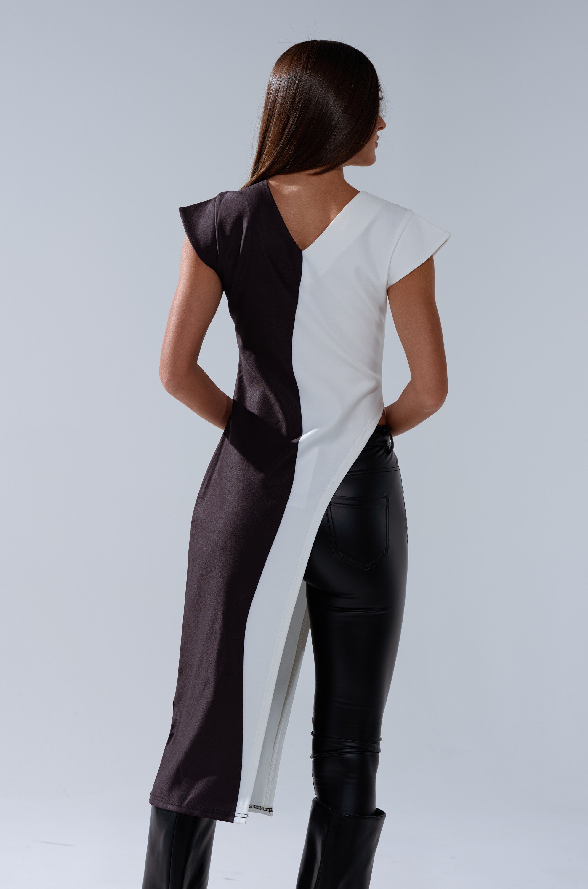 A woman with straight brown hair faces away, wearing the SILHOUETTE LONGLINE TEE styled as an asymmetrical dress over black leather pants and boots against a plain light background.
