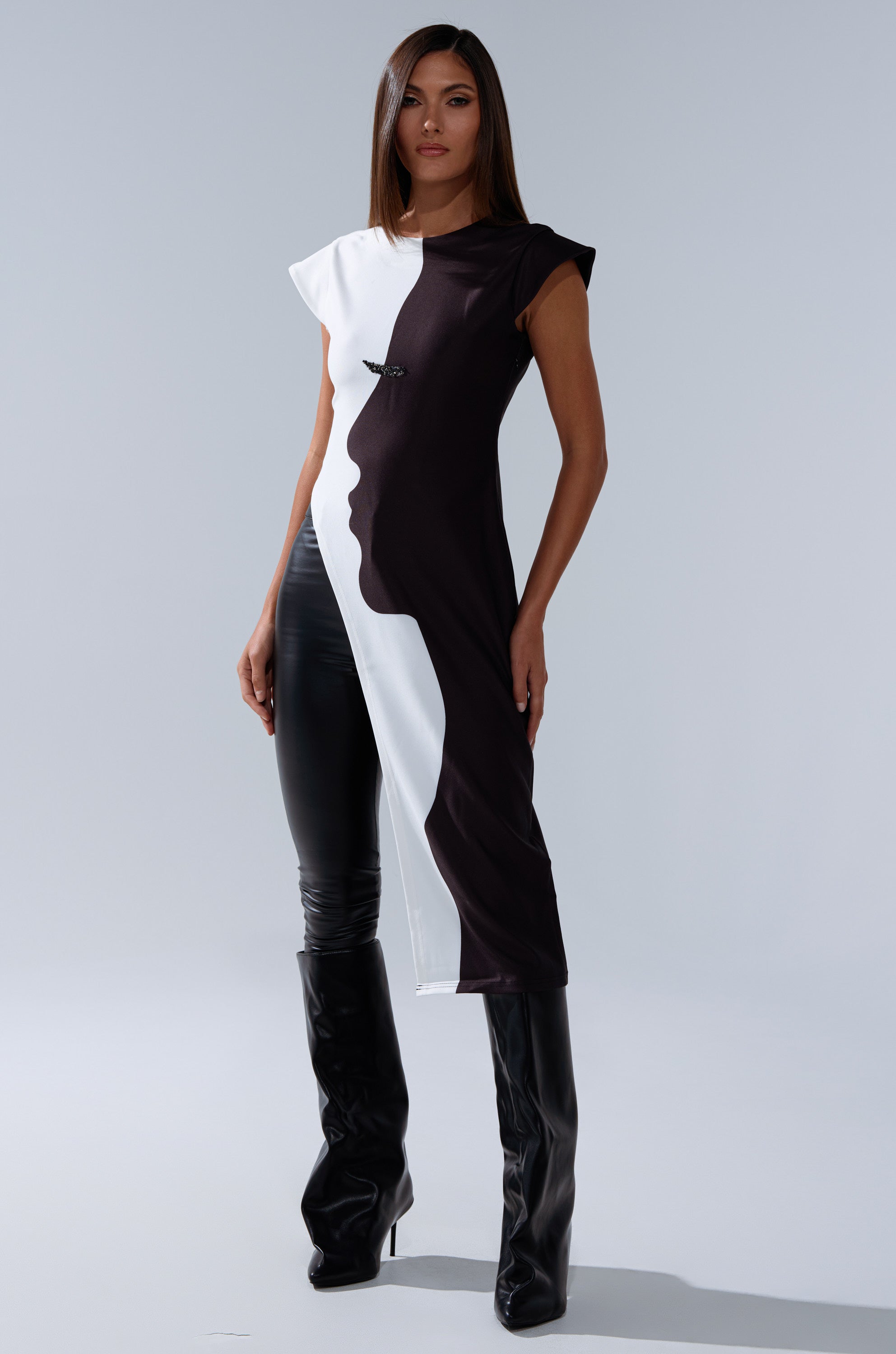 A woman stands confidently in the SILHOUETTE LONGLINE TEE, a striking two-tone top with an abstract face graphic, paired with black leather pants and knee-high boots against a plain light gray background.