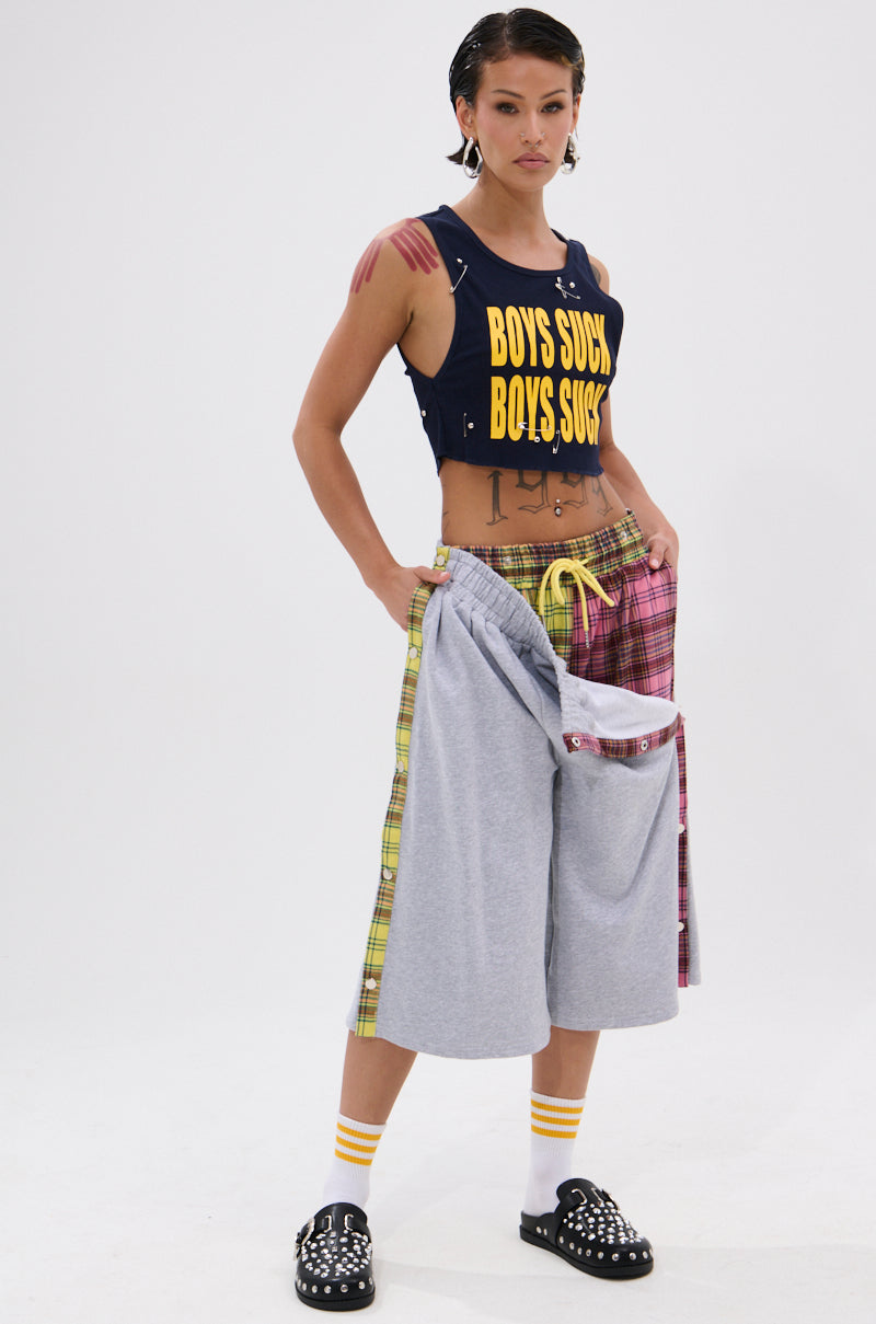A woman poses confidently in punk fashion, wearing the BOYS LIE DISTRESSED CROPPED TANK TOP, mismatched plaid and gray oversized shorts, striped socks, and studded black clogs against a white background. 3
