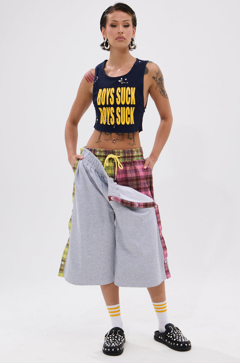 A person exudes punk edge in the BOYS LIE DISTRESSED CROPPED TANK TOP featuring bold yellow "BOYS SUCK" text, styled with patchwork shorts, striped socks, studded black shoes, visible tattoos, and short hair. 4