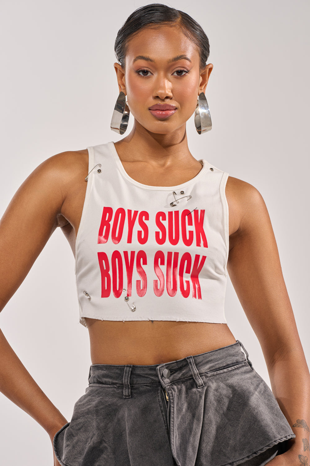 A woman wearing the BOYS LIE Distressed Cropped Tank in White with bold red lettering and high-waisted gray shorts stands confidently against a neutral background, accessorized with large hoop earrings. 0