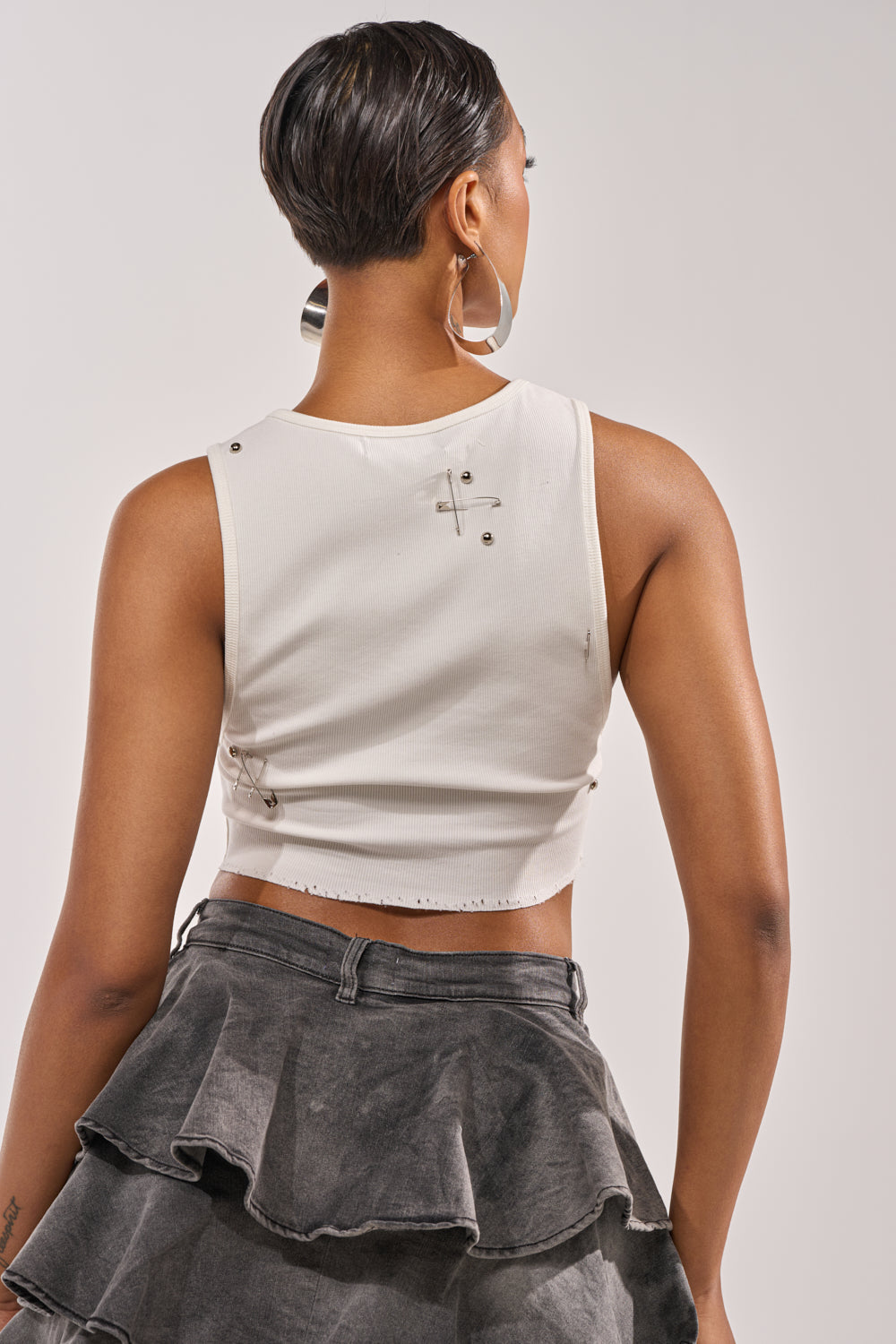 A person with short dark hair and large hoop earrings is seen from the back wearing the BOYS LIE DISTRESSED CROPPED TANK IN WHITE styled as a ribbed graphic crop top with metal studs and a black ruffled skirt, against a plain background. 3