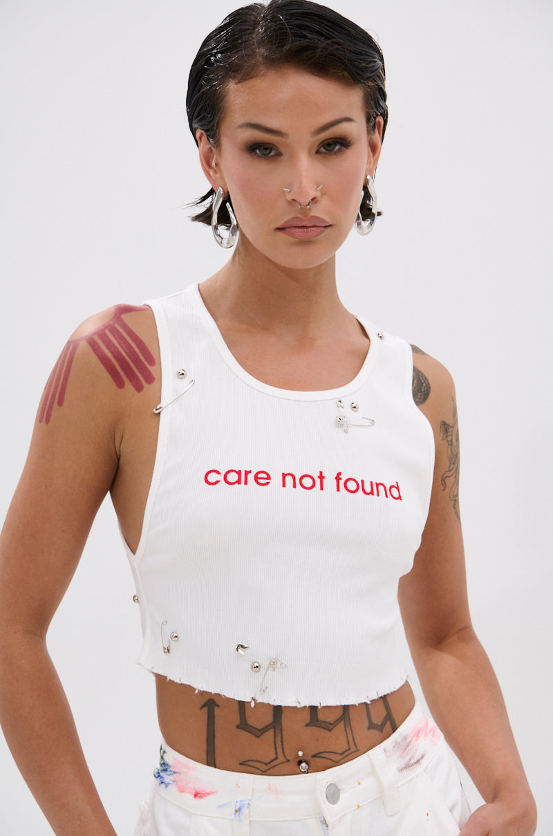 A person with short dark hair, hoop earrings, and facial piercings wears the NEVER CARED DISTRESSED CROPPED TANK TOP. Silver studs, safety pins, painted handprints, and tattoos complete their bold look. 0