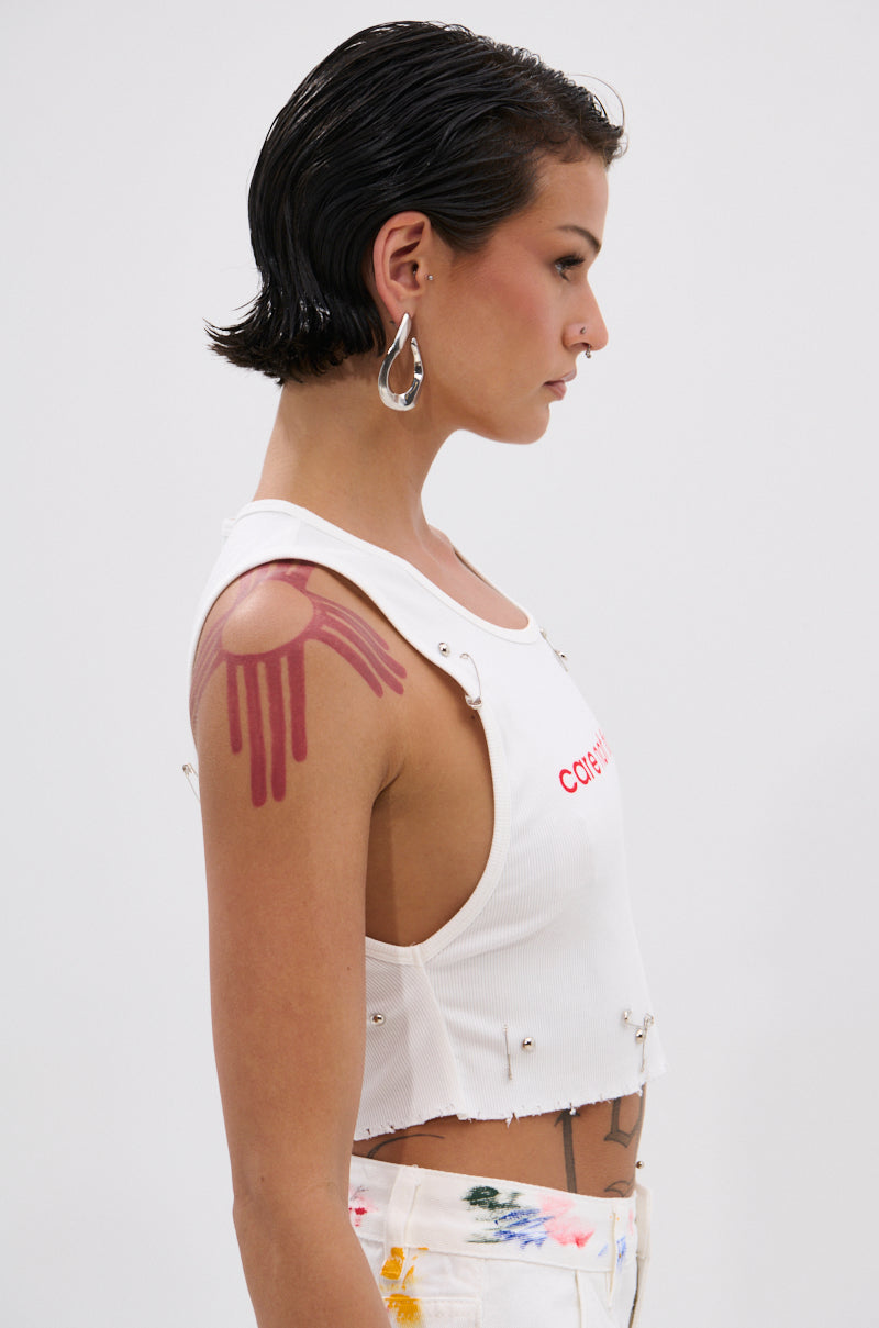 A person with short, slicked-back hair wears the NEVER CARED DISTRESSED CROPPED TANK TOP with silver studs, white paint-splattered pants, large hoop earrings, and a bold red abstract tattoo on their upper arm. 1