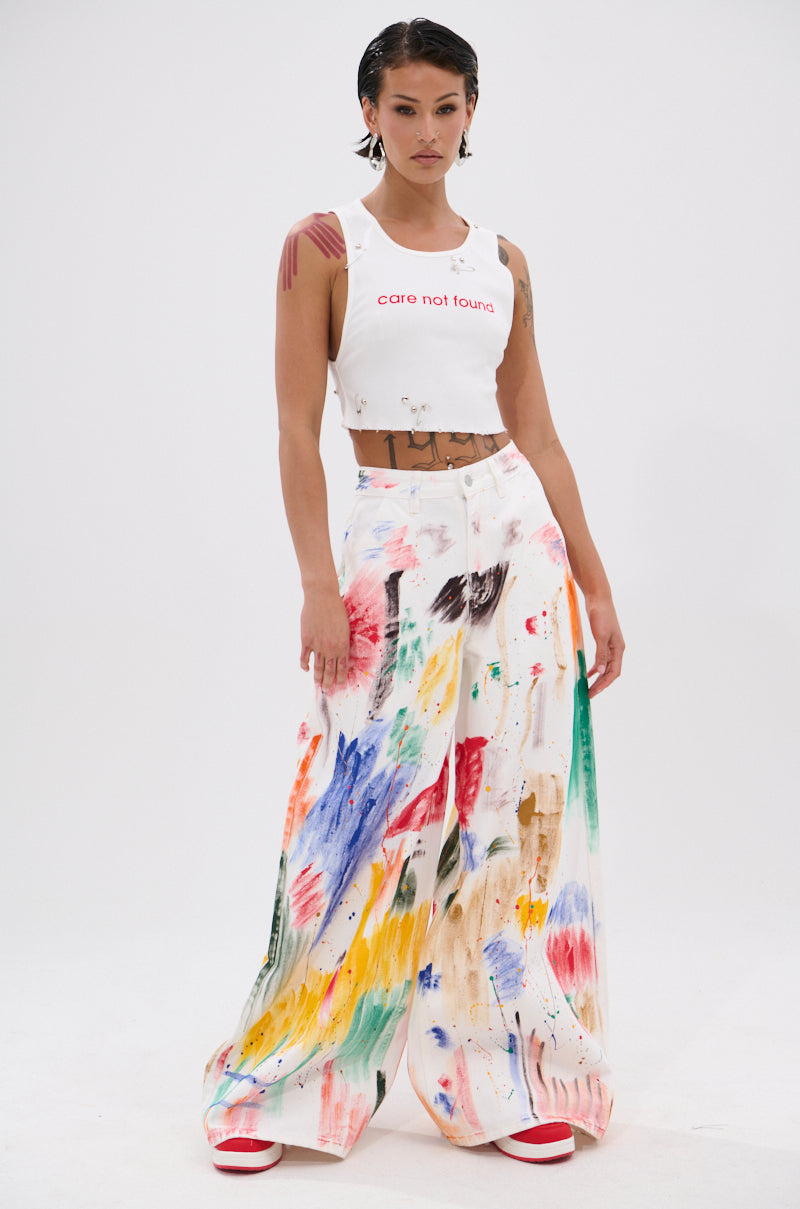 A woman with short, wet-look hair stands against a plain background wearing the NEVER CARED DISTRESSED CROPPED TANK TOP, featuring "care not found" text, paint-splattered wide-leg pants, and red-and-white shoes. 4