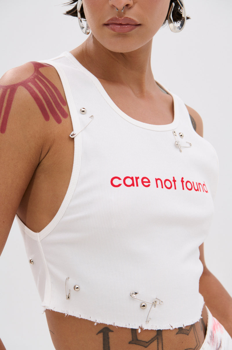 A person wears the NEVER CARED DISTRESSED CROPPED TANK TOP—a curve-hugging, ribbed crop with frayed edges, safety pins, and bold red “care not found” lettering—plus large hoop earrings, a nose ring, and a red shoulder tattoo. 5