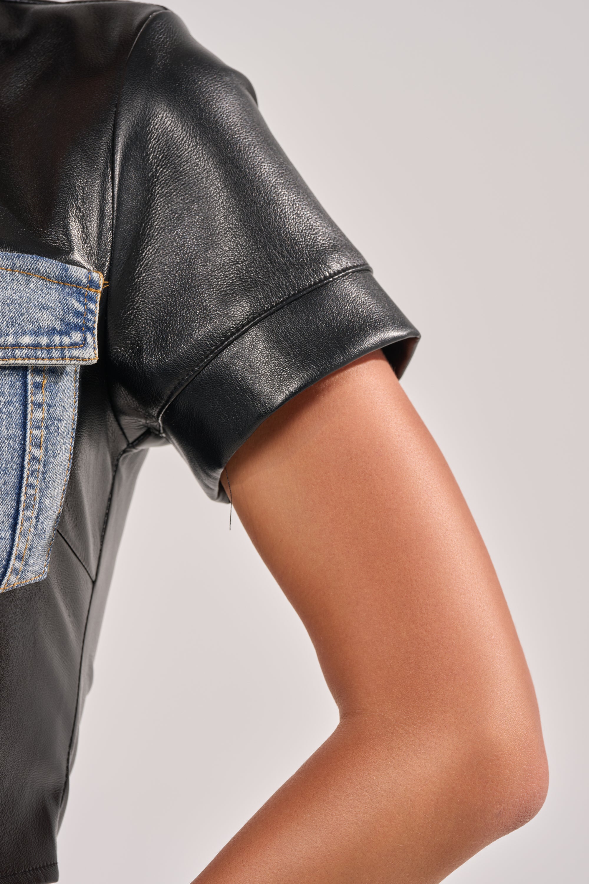 Close-up of a bent arm wearing the MIXED VIBES FAUX LEATHER SHORT SLEEVE BUTTON DOWN, a black faux leather cropped style with denim pocket detail. Set against a plain, light background. 8