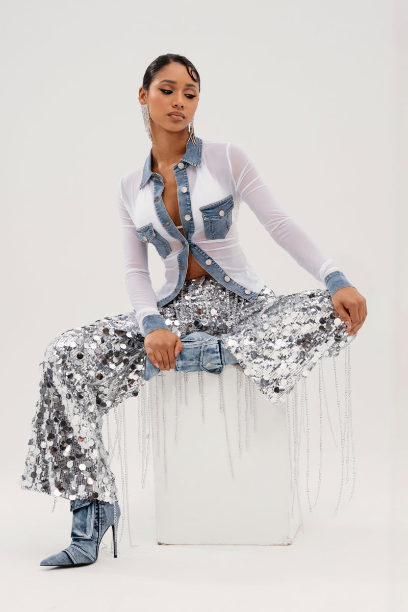 Confidently posing on a white block, the person flaunts the DIFFERENT LOVE MESH BUTTON DOWN paired with high-waisted silver sequin pants with fringe. Metallic boots and large hoop earrings complete their sleek style, set against a plain background.