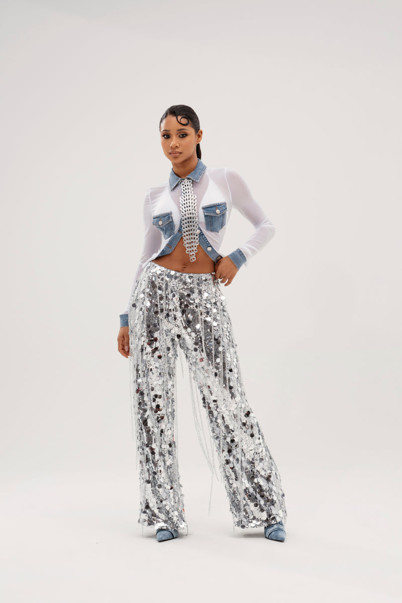 A person confidently wears the DIFFERENT LOVE MESH BUTTON DOWN, featuring denim accents and a tie, paired with sparkling silver sequin pants against a plain white background.
