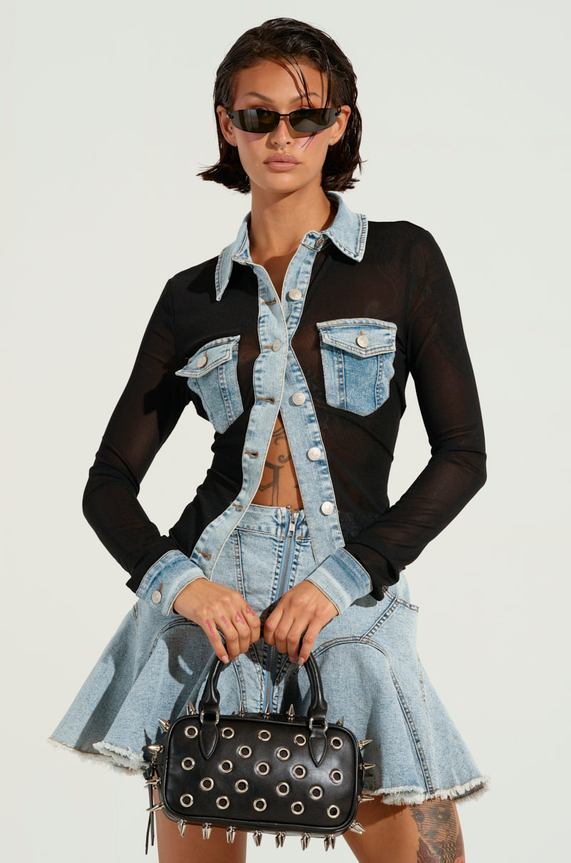 A person in a denim skirt and jacket with sheer sleeves stands confidently holding a black handbag adorned with eyelet details. Exuding mixed vibes, they wear sunglasses and have short, dark hair, perfectly capturing the essence of a MIXED VIBES BUTTON DOWN BLOUSE moment.