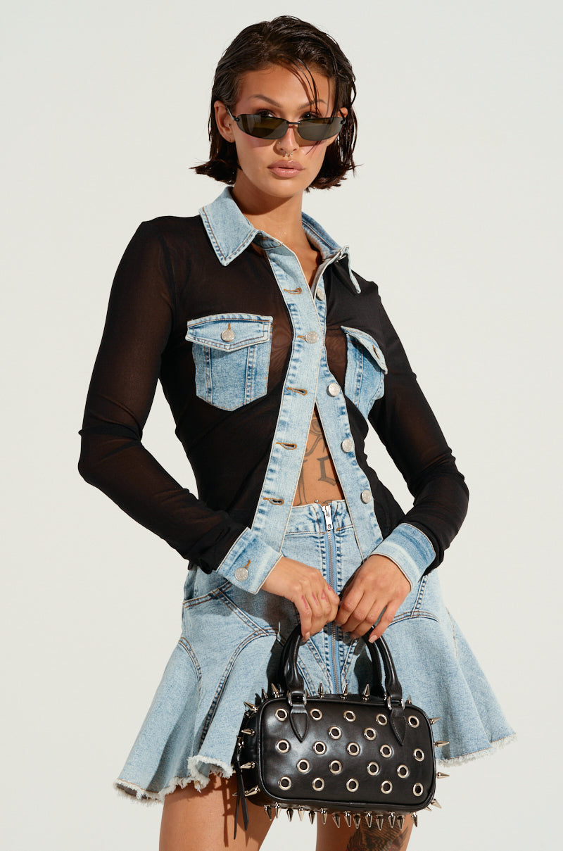 With mixed vibes, a person with short hair and sunglasses confidently poses in a denim skirt and the AKIRA Label "Mixed Vibes Button Down Blouse," featuring denim pockets. They hold a black handbag adorned with metal eyelets and spike details against a plain background.