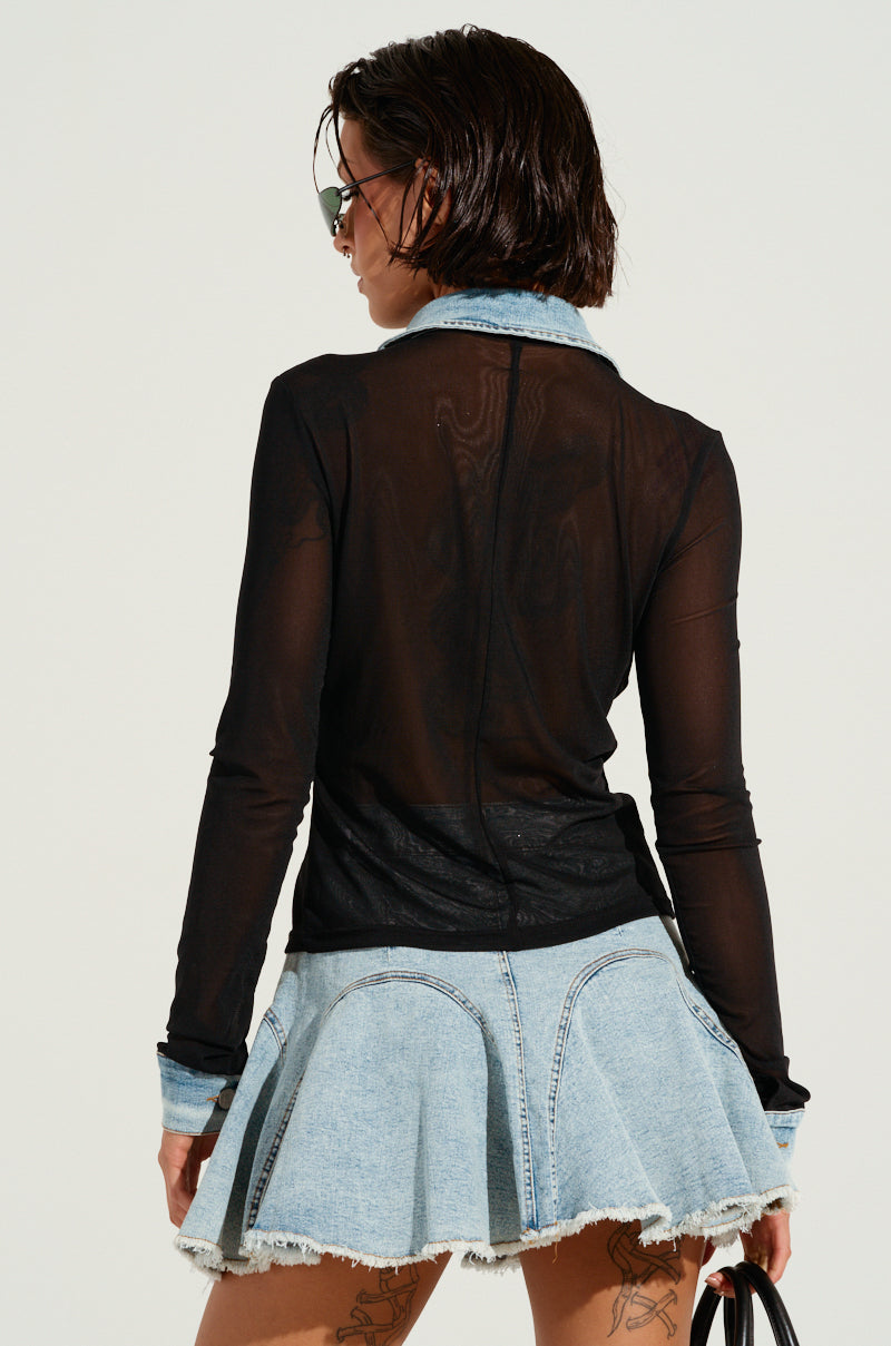 A person with short dark hair is rocking oval sunglasses and a sheer mesh top that brings to mind the MIXED VIBES BUTTON DOWN BLOUSE, paired with a denim mini skirt featuring a frayed hem. They are turned slightly away, holding a black handbag and exuding mixed vibes. Intricate tattoo designs adorn their legs.