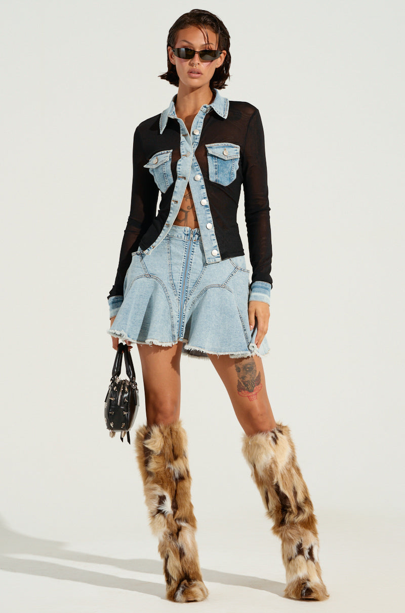 A person wearing sunglasses, a MIXED VIBES BUTTON DOWN BLOUSE with denim accents, a short denim skirt, and furry knee-high boots. They are holding a small handbag and standing against a plain background, exuding mixed vibes.