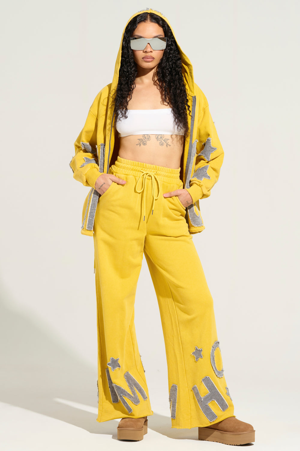 A person confidently stands in a striking yellow AKIRA TELL HER ABOUT ME JOGGER tracksuit, featuring gray star details and a hood. The high-rise design beautifully complements their long curly hair, reflective sunglasses, white crop top, and platform shoes, resulting in a modern and audacious appearance.