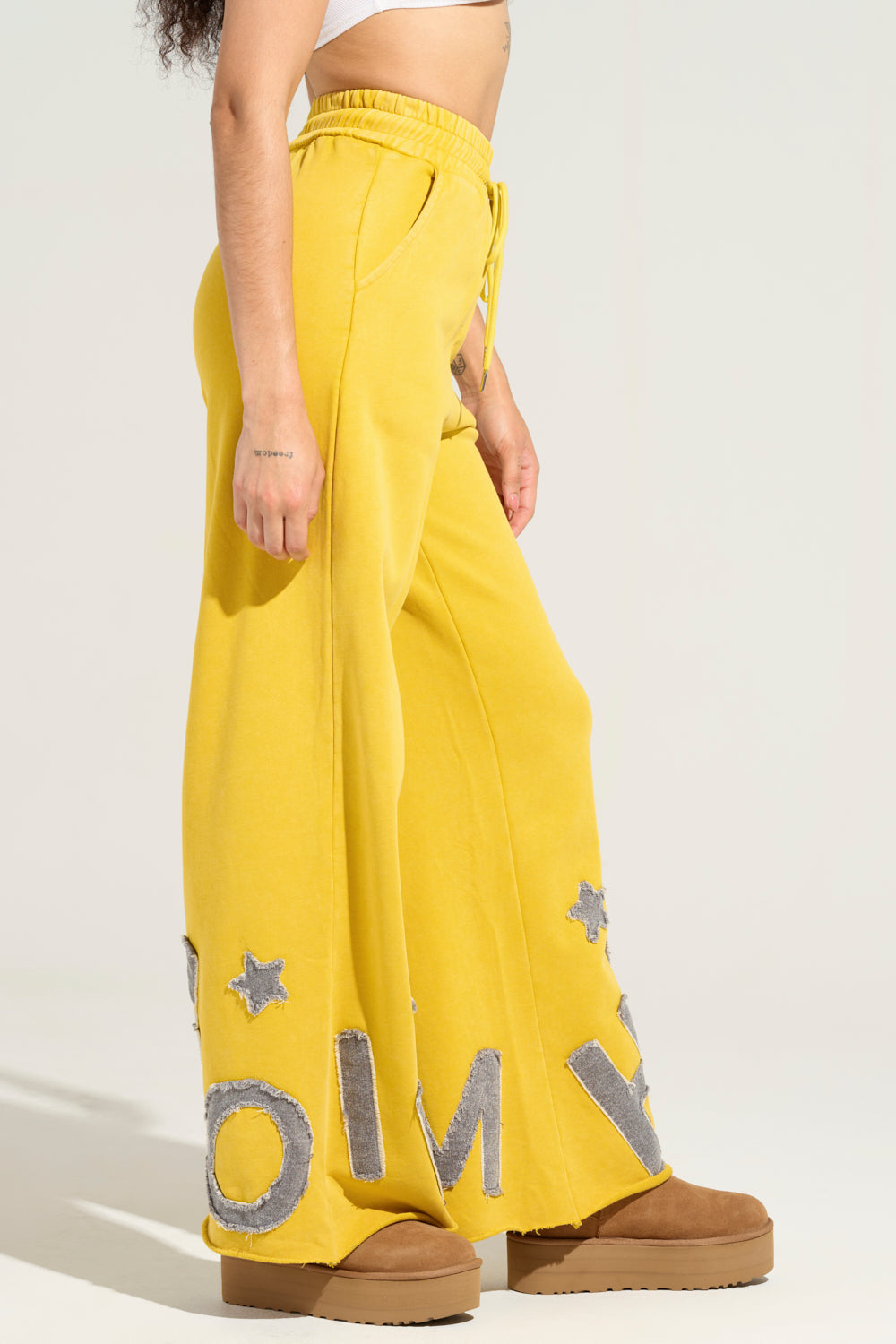 Wearing the TELL HER ABOUT ME JOGGER, featuring yellow wide-leg pants adorned with large gray letters and stars at the bottom, reminiscent of the AKIRA Label Jogger style, the person completes their outfit with brown boots and a white top against a plain background. A small tattoo is visible on their right arm.