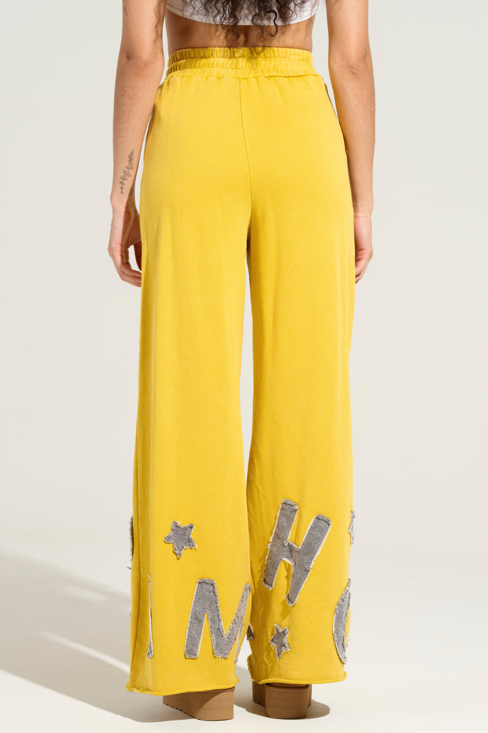 The individual is styled in TELL HER ABOUT ME JOGGER pants by AKIRA, which boast a vibrant yellow hue and are embellished with silver stars and letters near the bottom. These high-rise wide-leg joggers are made from a stretchy cotton blend and feature an elastic waistband as they face away from the camera.
