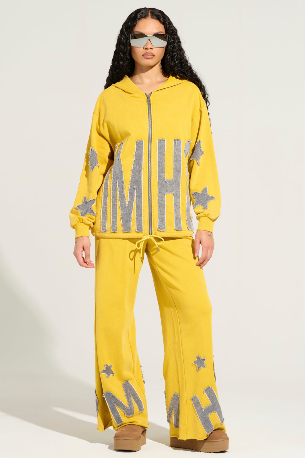 A woman wears a bright yellow TELL HER ABOUT ME JOGGER, crafted from a stretchy cotton blend and adorned with large silver "M" and "H" letters and star patterns. Complementing her long dark hair, sunglasses, and brown wedge shoes, she stands confidently against a plain light background.