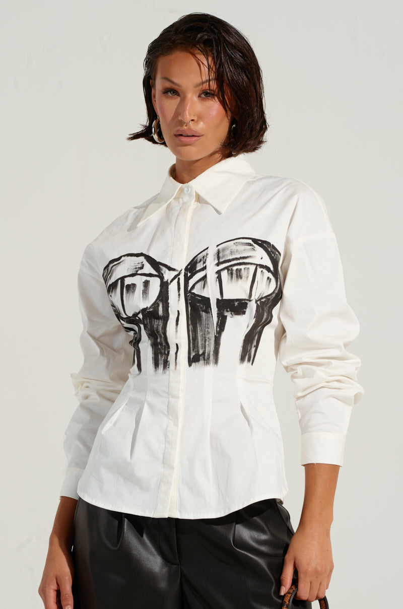 A woman poses in the BAD FOR YOU BUTTON DOWN BLOUSE, highlighted by a bustier-inspired graphic. Her short, wet-look hair complements her edgy style, perfectly paired with black leather pants. The plain, light-colored background enhances the blouse's bold design.