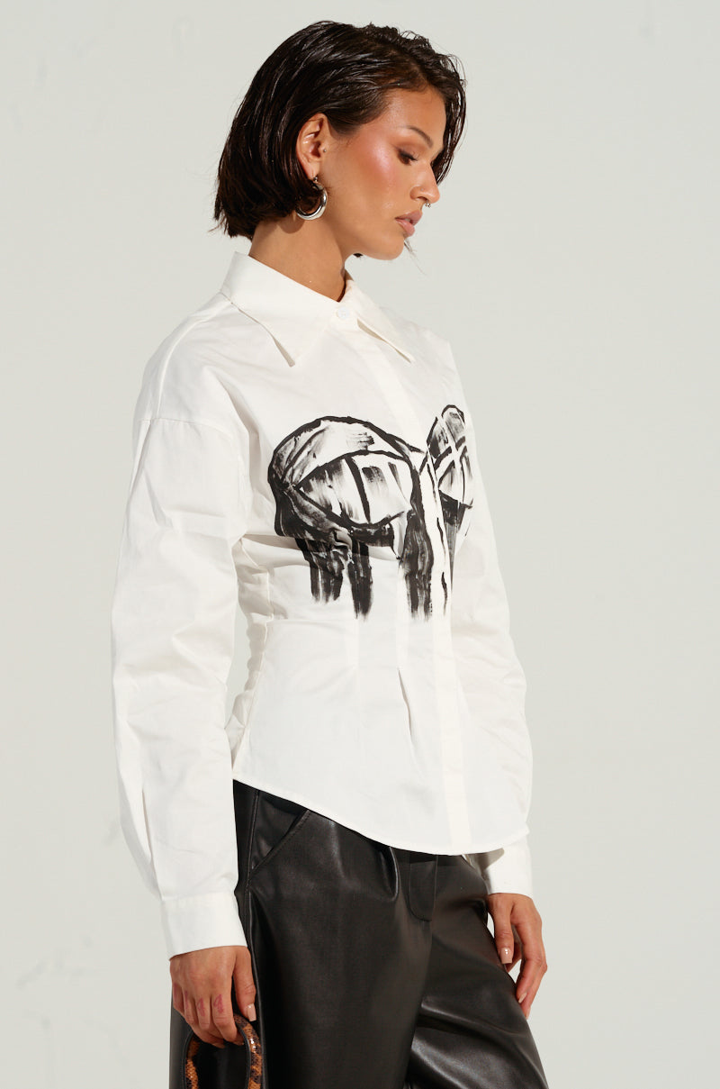 A person with short dark hair wears the BAD FOR YOU BUTTON DOWN BLOUSE in white, showing off its bold, bustier-inspired graphic on the front. They pair it with black leather pants and have a neutral expression while standing against a plain background.