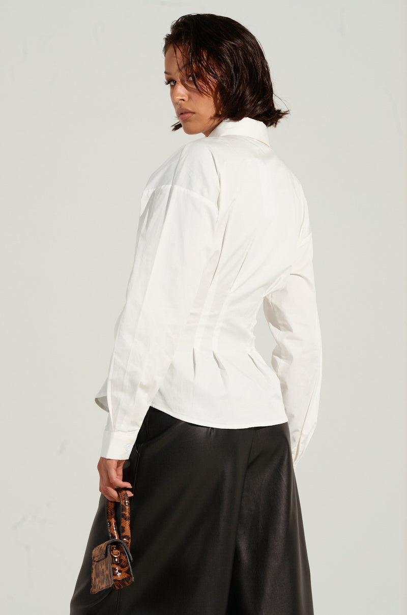 A person with short, dark hair is wearing the BAD FOR YOU BUTTON DOWN BLOUSE in white, styled like a fitted bustier-inspired graphic shirt, along with black leather pants. They are holding a small, brown patterned bag and looking over their shoulder against a plain background.
